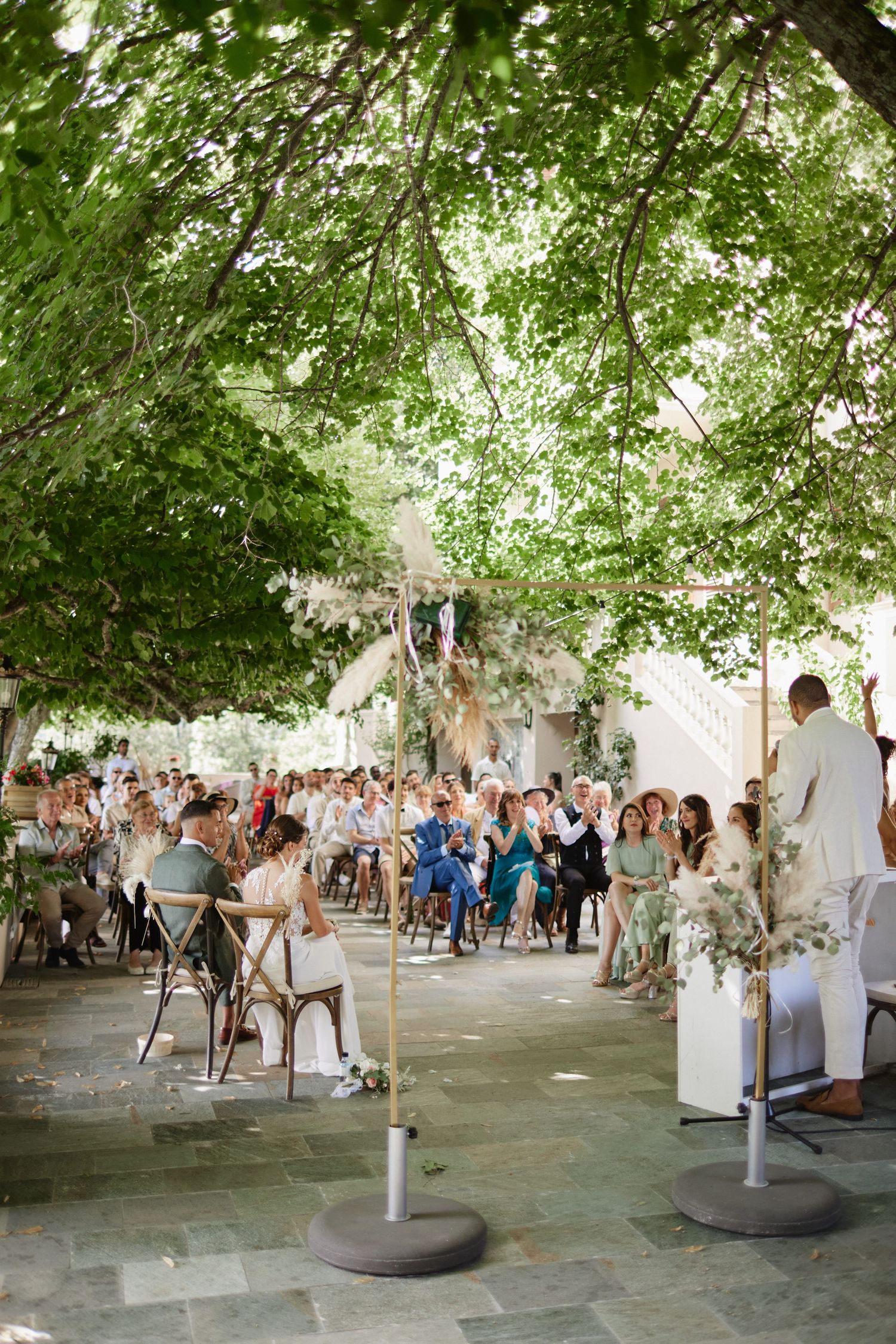 Wide view of outdoor wedding ceremony with guests seated under canopy of green trees.