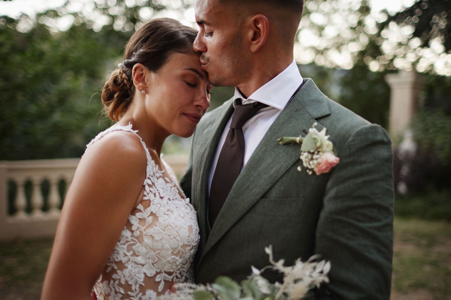 Groom in green suit tenderly kisses brides forehead as she closes her eyes during outdoor wedding portrait.