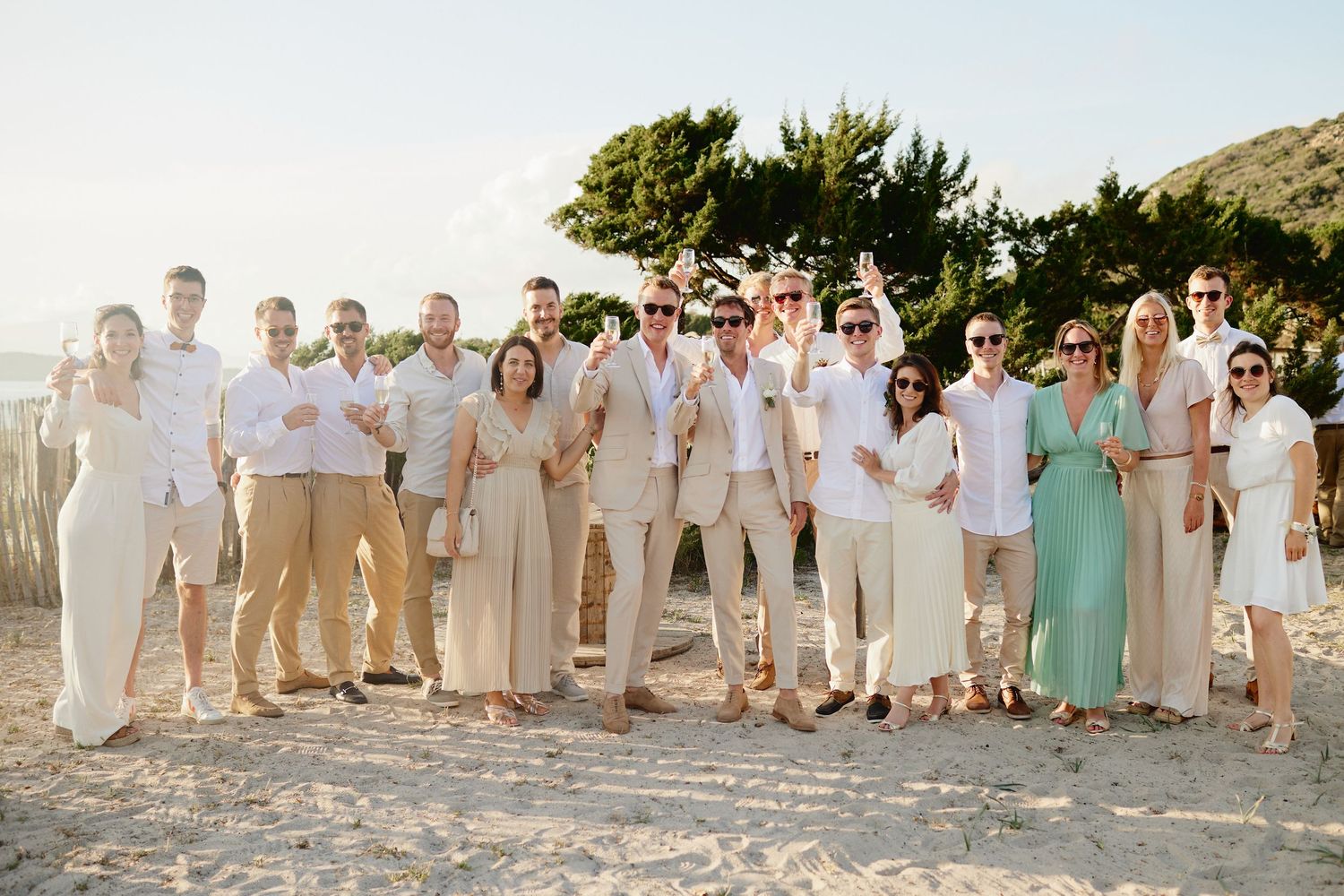 Large wedding party group photo on a sunny beach with guests in white and neutral tones.