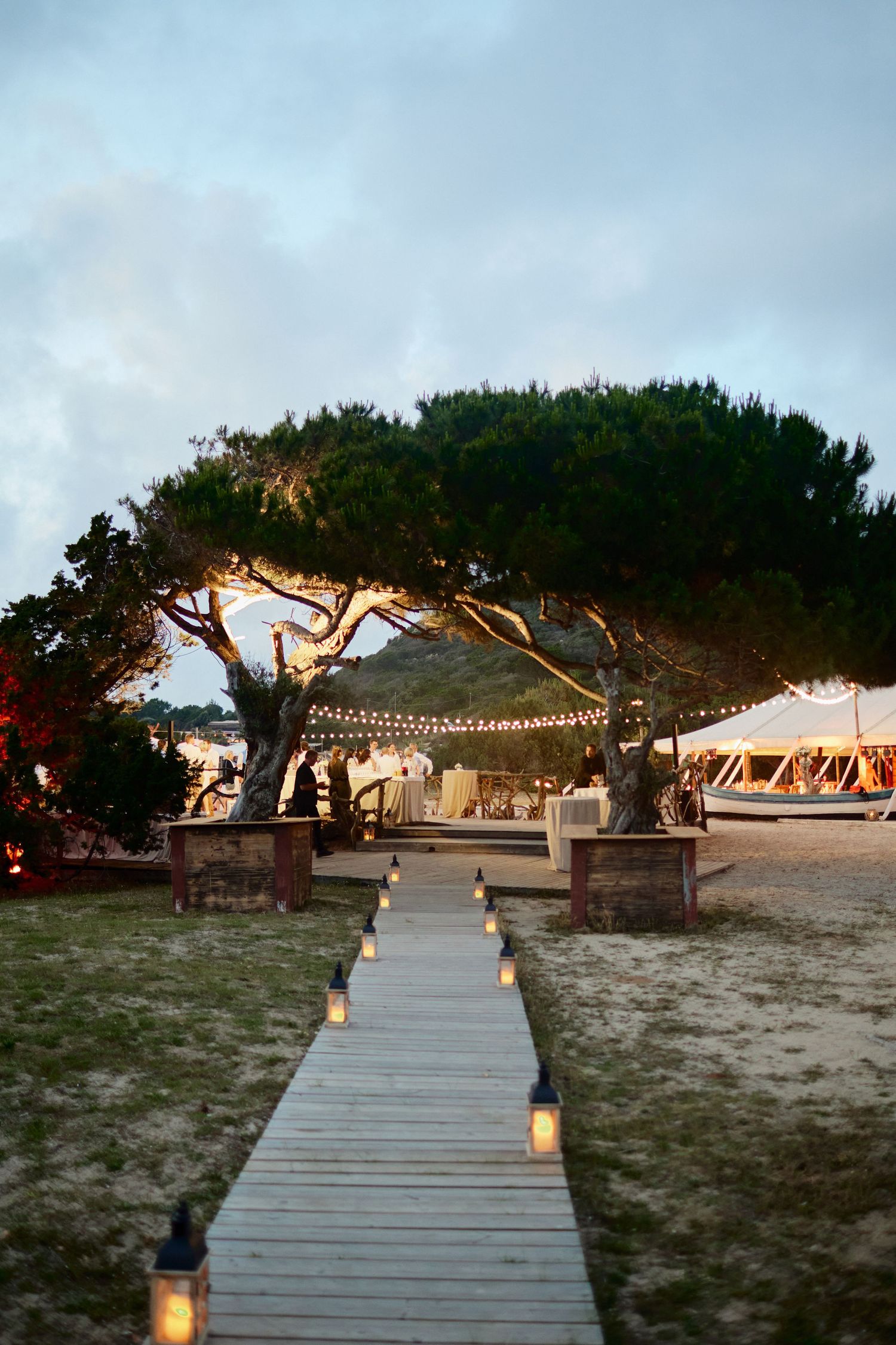 A candlelit wooden boardwalk leads to an elegant outdoor beach wedding reception tent at dusk.