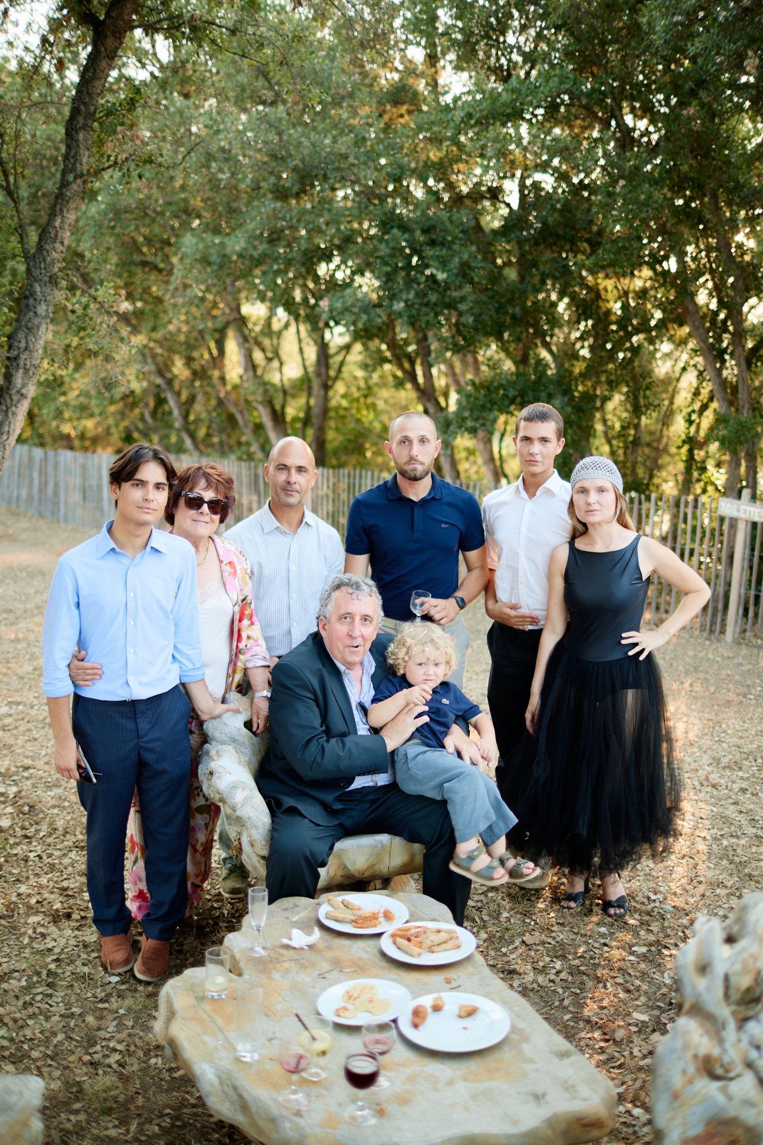 Family group portrait gathered around stone table in wooded outdoor setting at wedding.