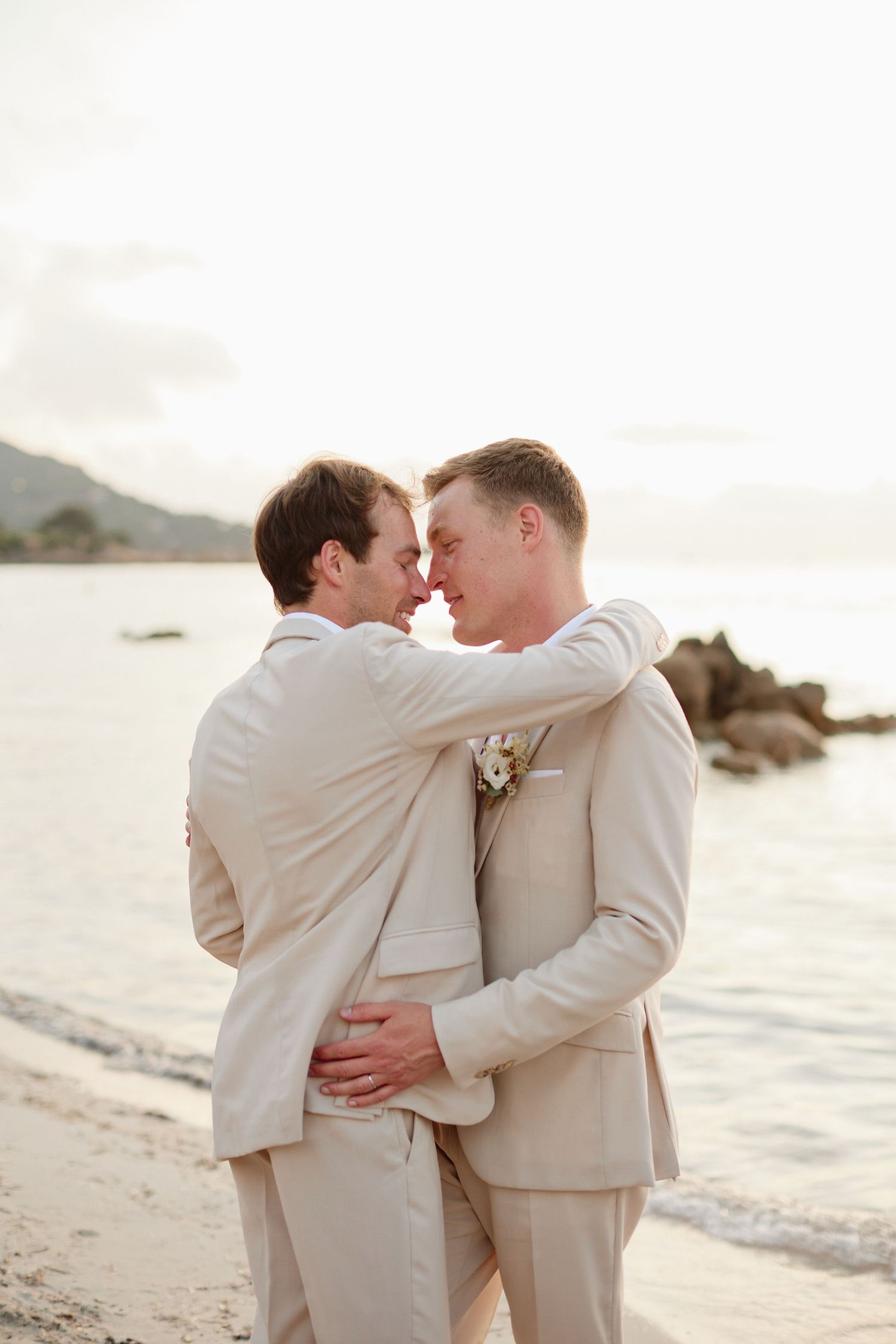 Two grooms in matching beige suits sharing an intimate moment on a beach at golden hour.