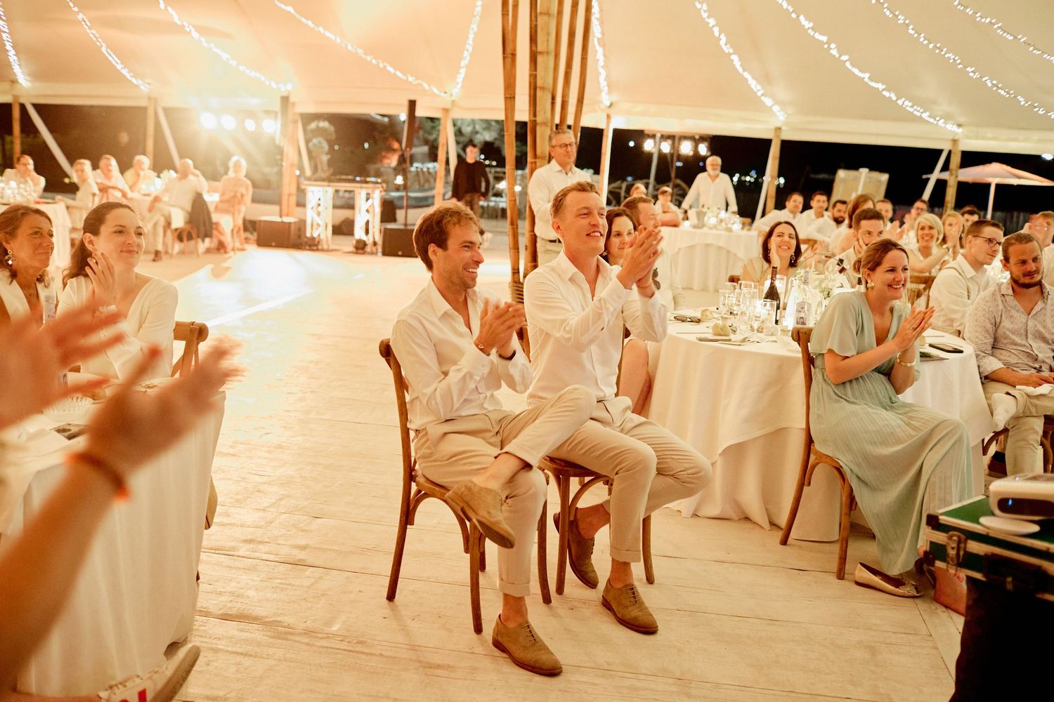 Two grooms clapping and smiling with wedding guests seated at tables under a warmly lit reception tent.