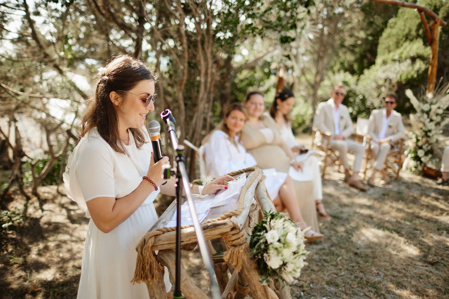 A woman in white speaks at a microphone podium during an outdoor wedding ceremony.
