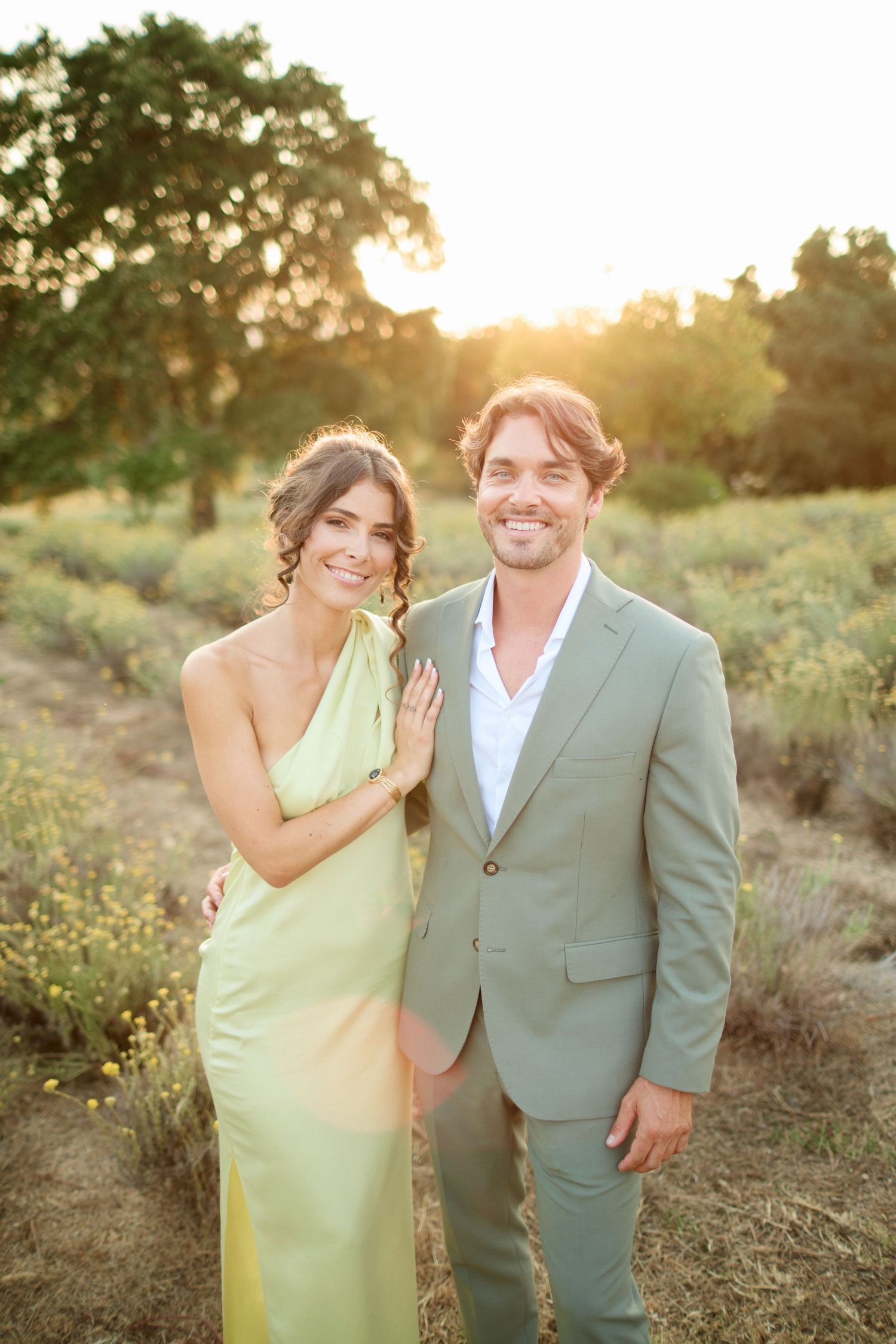 Couple poses together in golden sunset light, woman in green gown and man in sage suit.