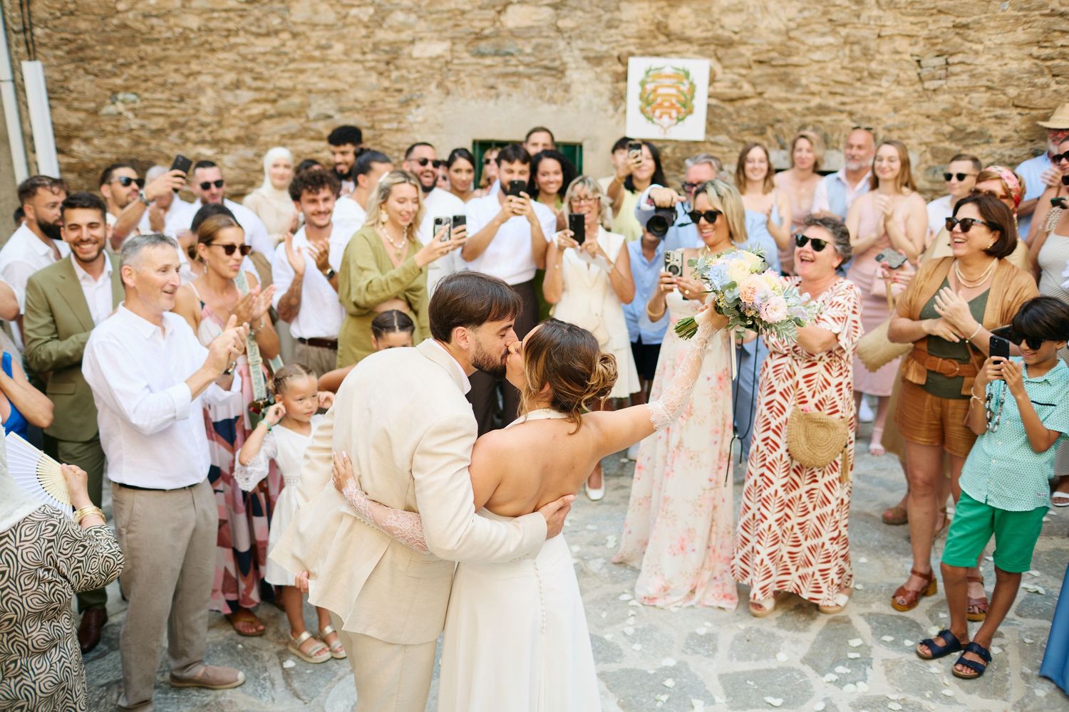 Newlyweds kiss surrounded by cheering guests outside rustic stone building after ceremony.