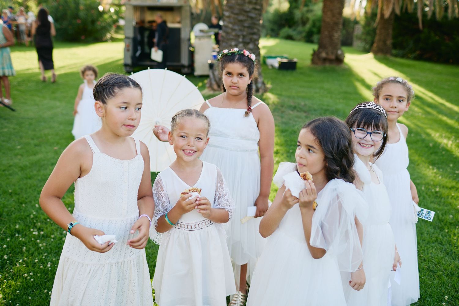 Group of young flower girls dressed in white gather on lawn holding parasols at an outdoor wedding.