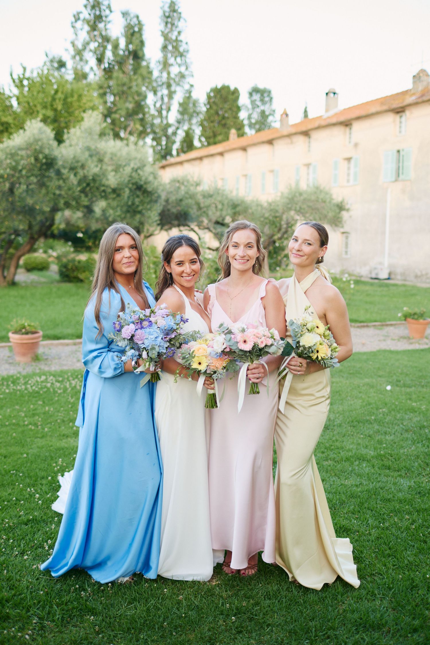 Four women in pastel bridesmaid dresses holding floral bouquets posing together on lawn at French estate.