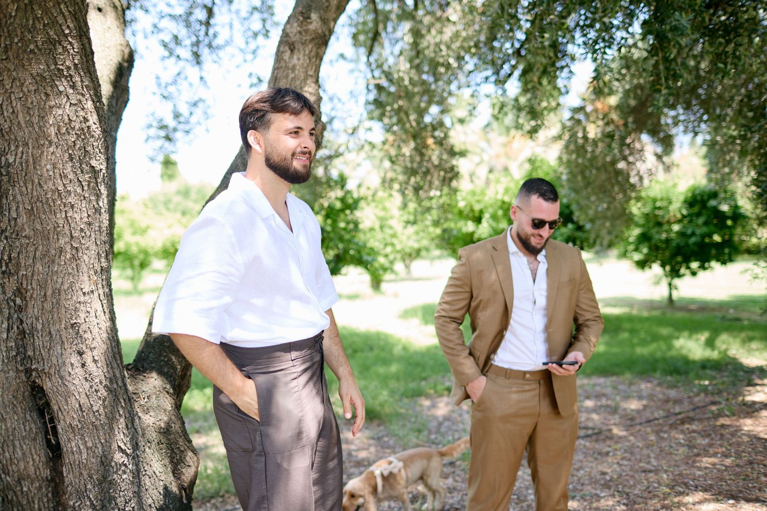 Two well-dressed men in a sunny garden, one in white shirt leaning against a tree with a dog.