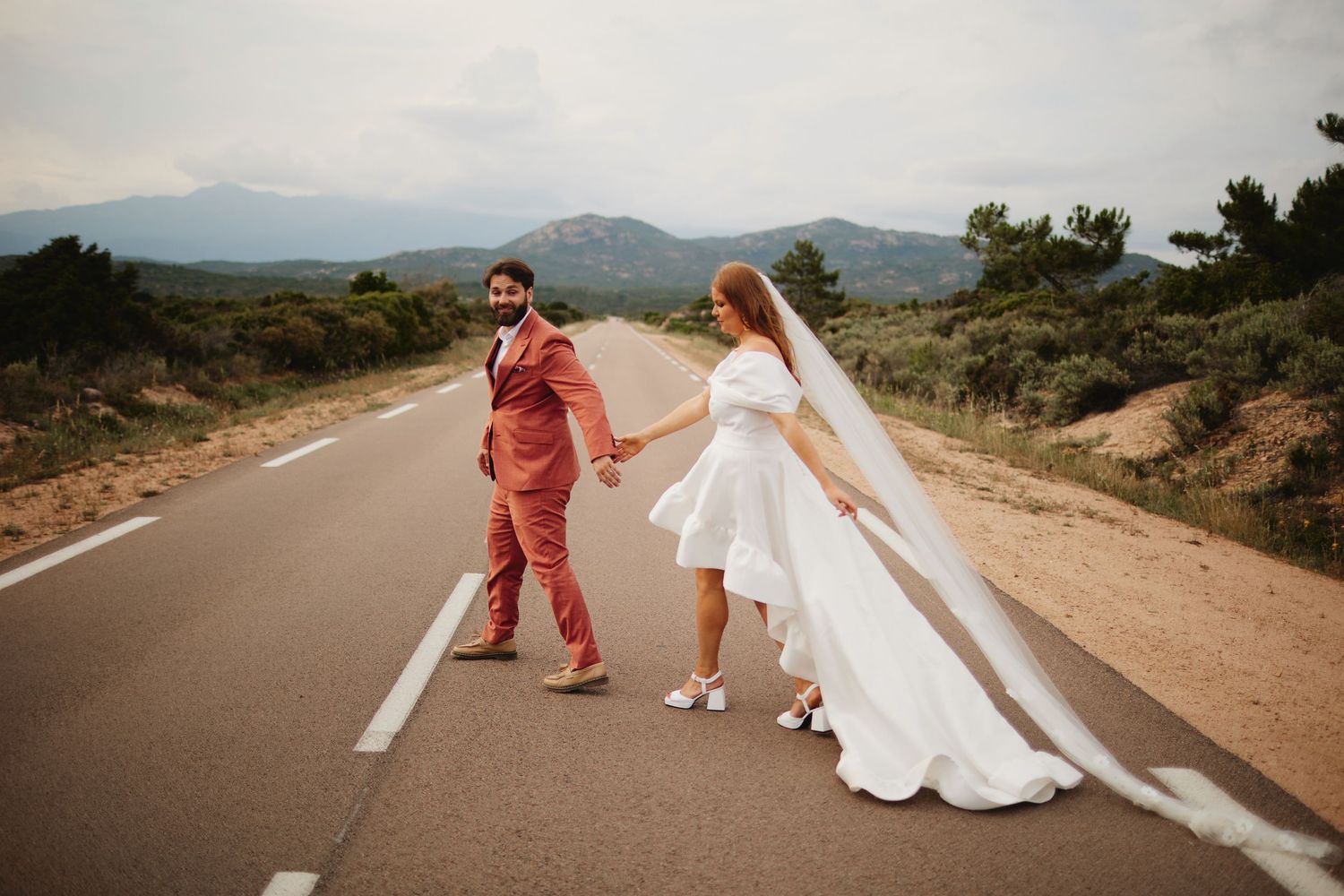 Newlyweds hold hands walking down an empty road with mountains and greenery in the background.