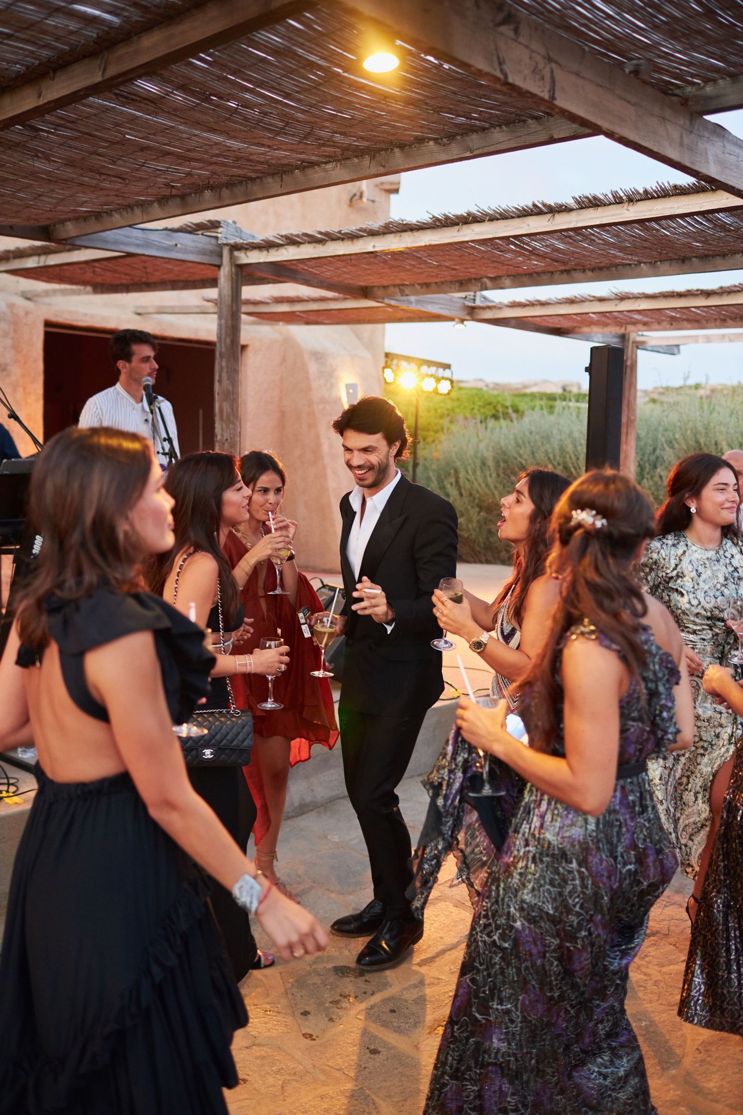 Guests in formal attire dance and celebrate at an outdoor wedding reception under pergola.