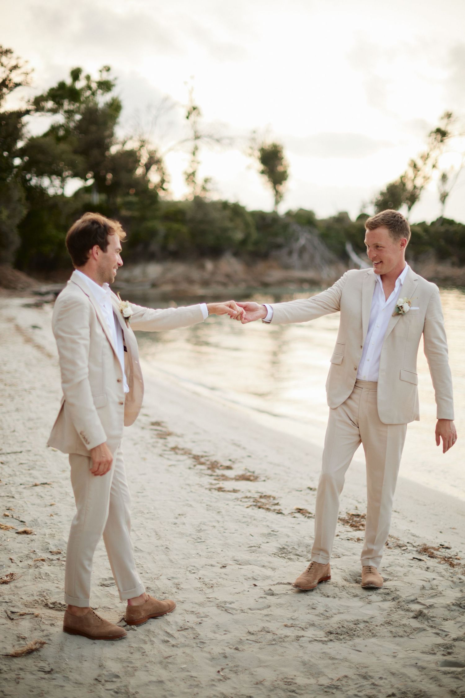 Two grooms in matching beige linen suits hold hands on a sandy beach at sunset.