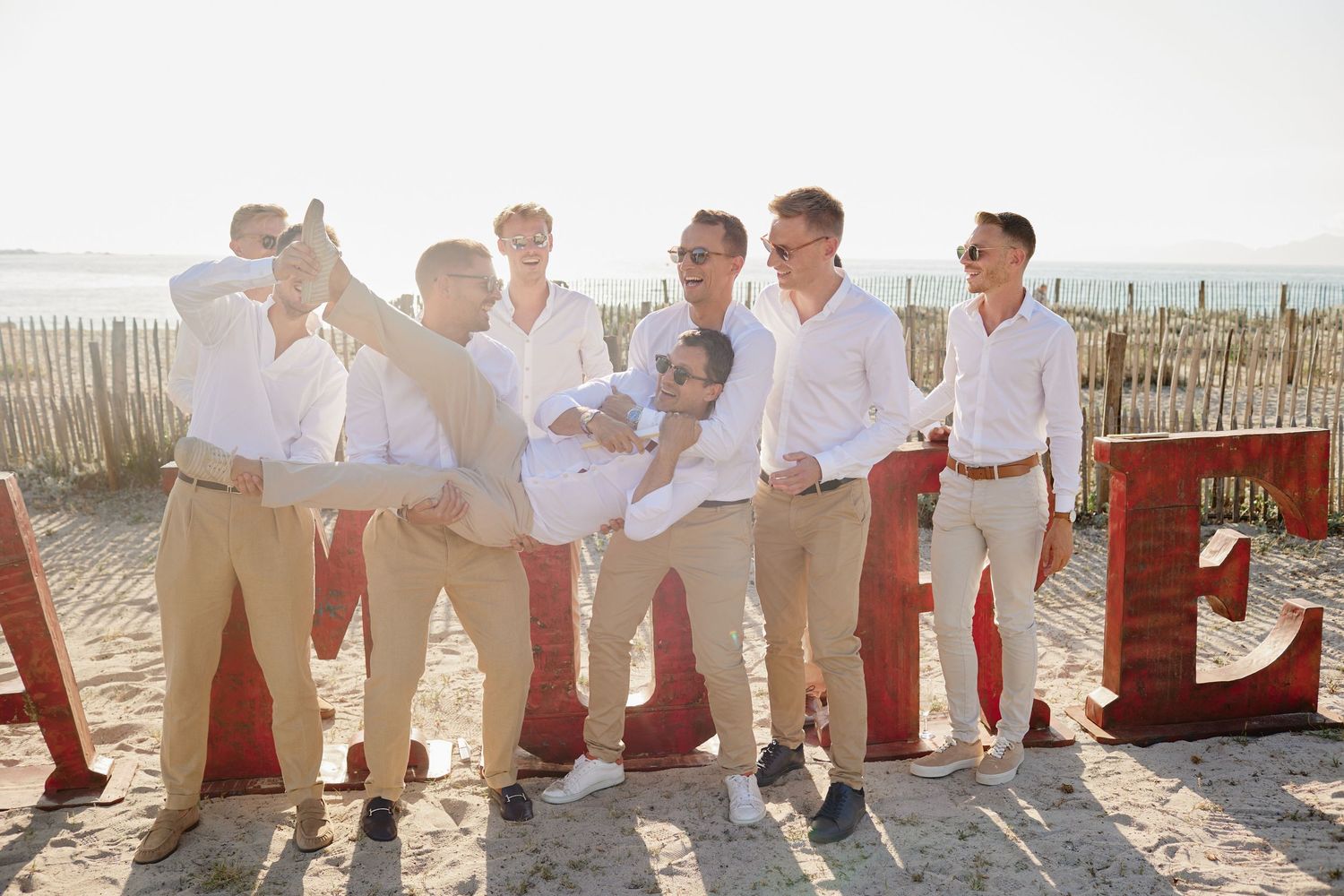 Groomsmen in white shirts and khaki pants posing and laughing on a sandy beach at a wedding.
