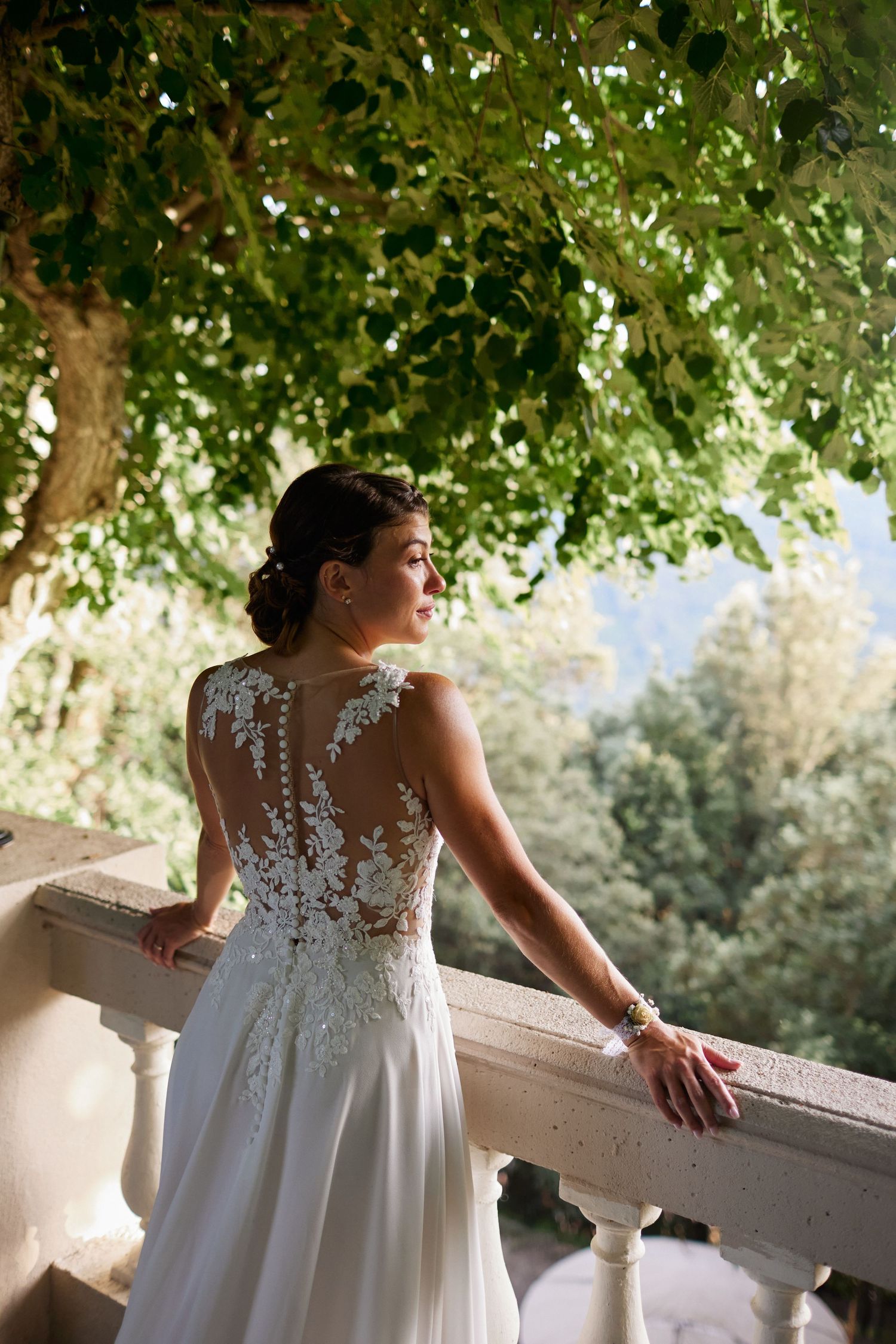 Bride in lace-back wedding gown stands on balcony surrounded by lush green trees.