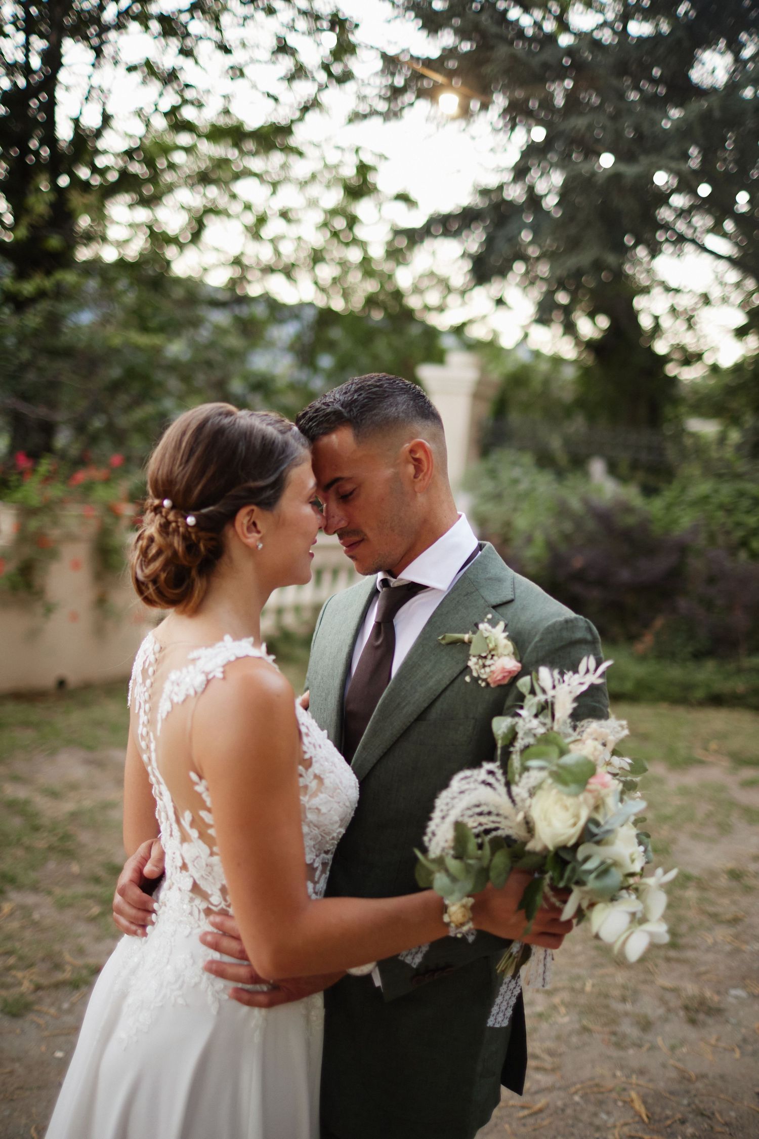 Newlyweds share a tender close moment in garden, bride in backless lace dress, groom in olive green suit.