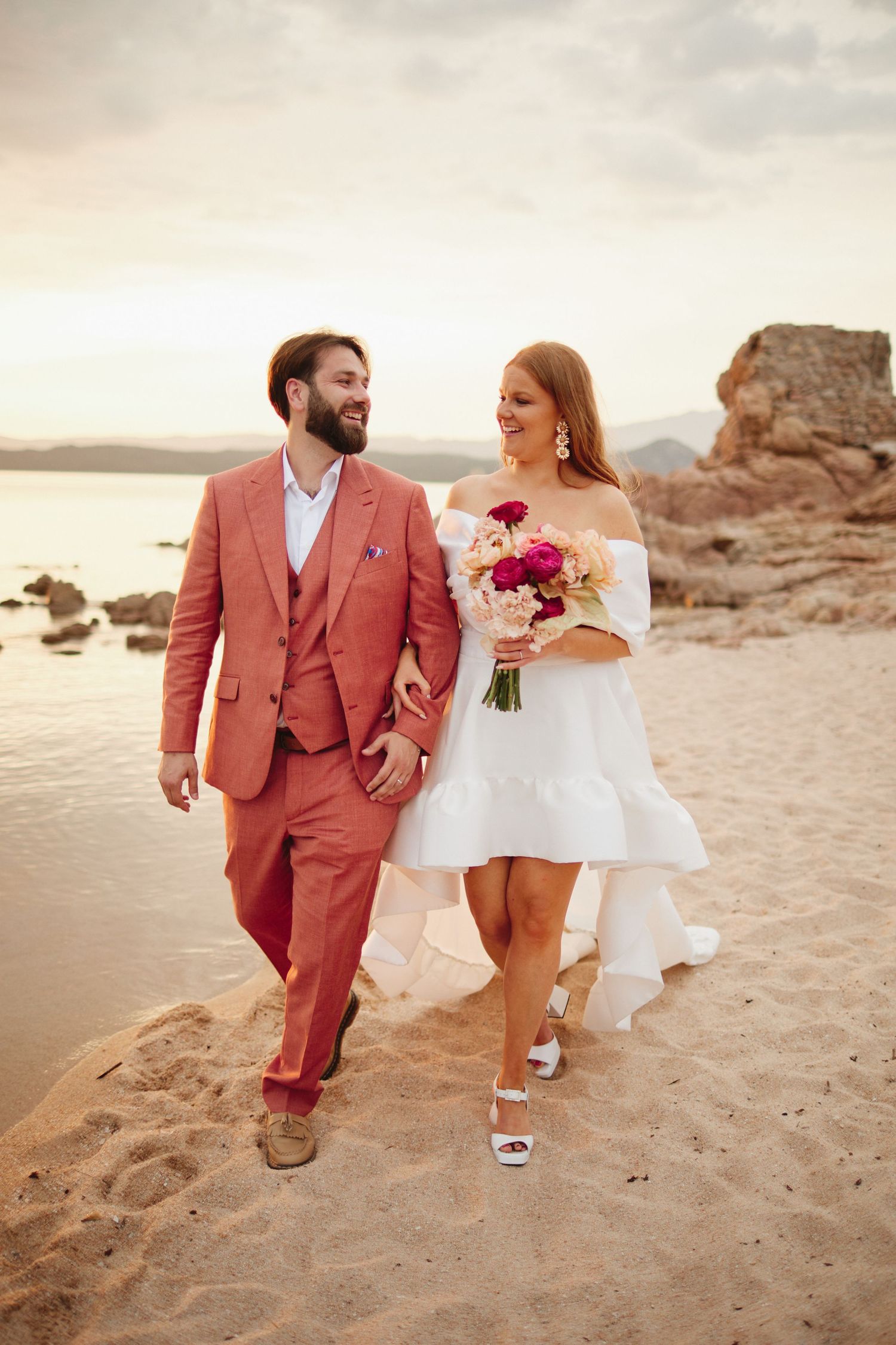 Smiling bride and groom in pink suit walking hand-in-hand on a sandy beach at golden sunset.