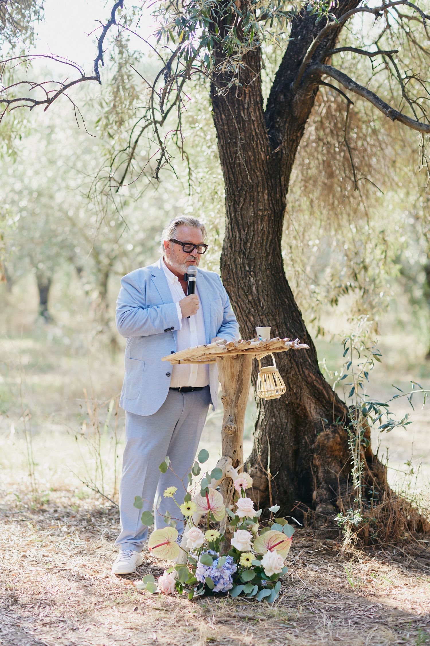 Officiant in light blue suit speaks at rustic wooden altar decorated with flowers under olive tree.