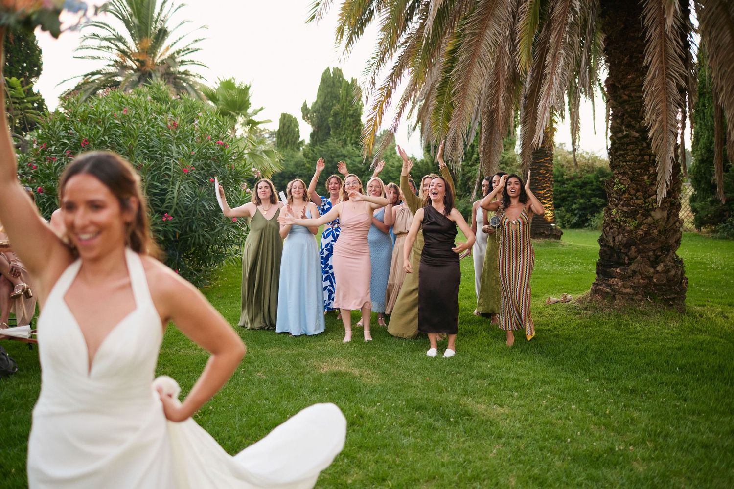 Bride tossing bouquet to laughing and cheering bridesmaids and guests at outdoor garden wedding celebration.