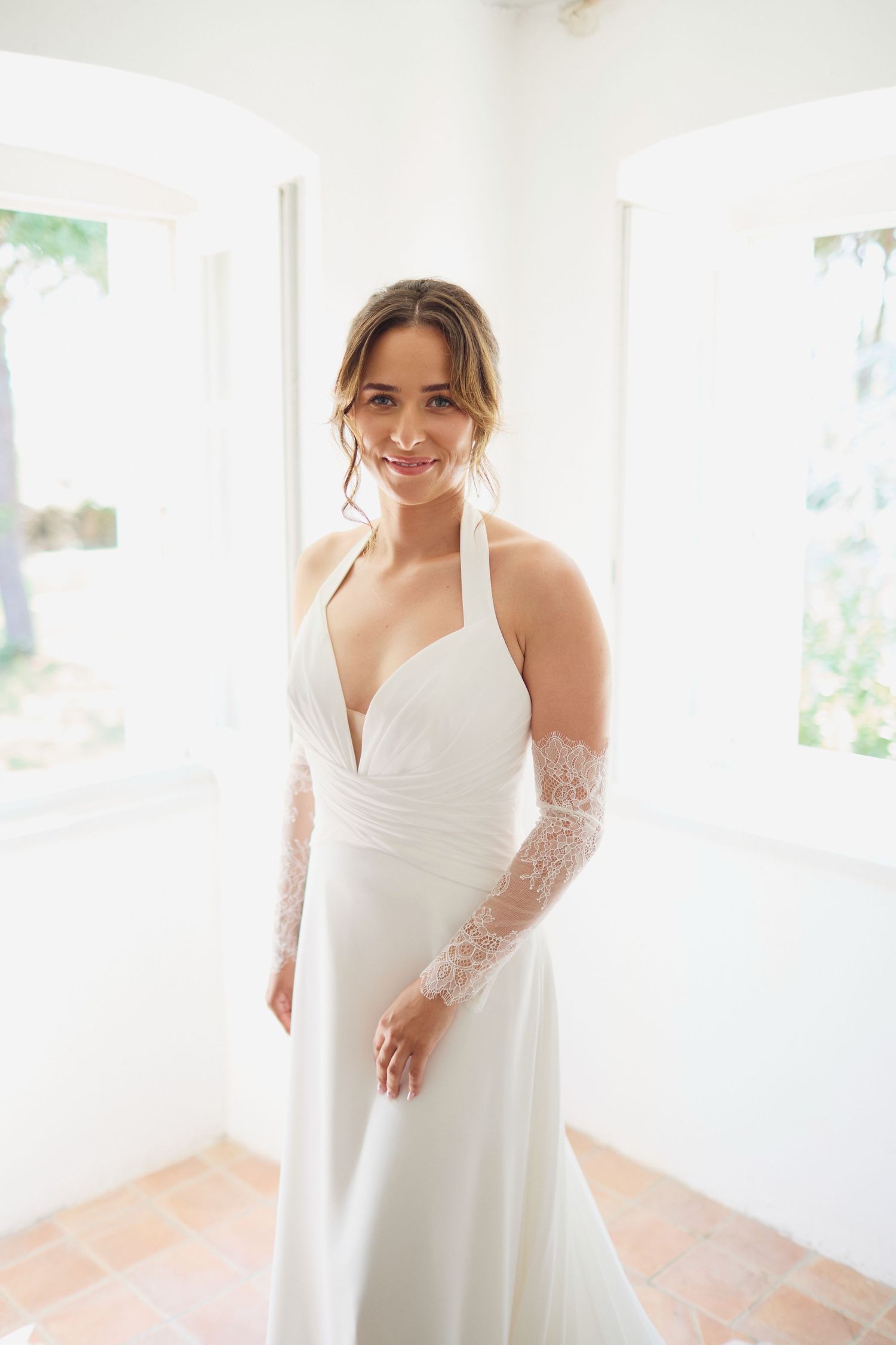 Smiling bride in white halter wedding gown with lace sleeves posing in bright doorway.