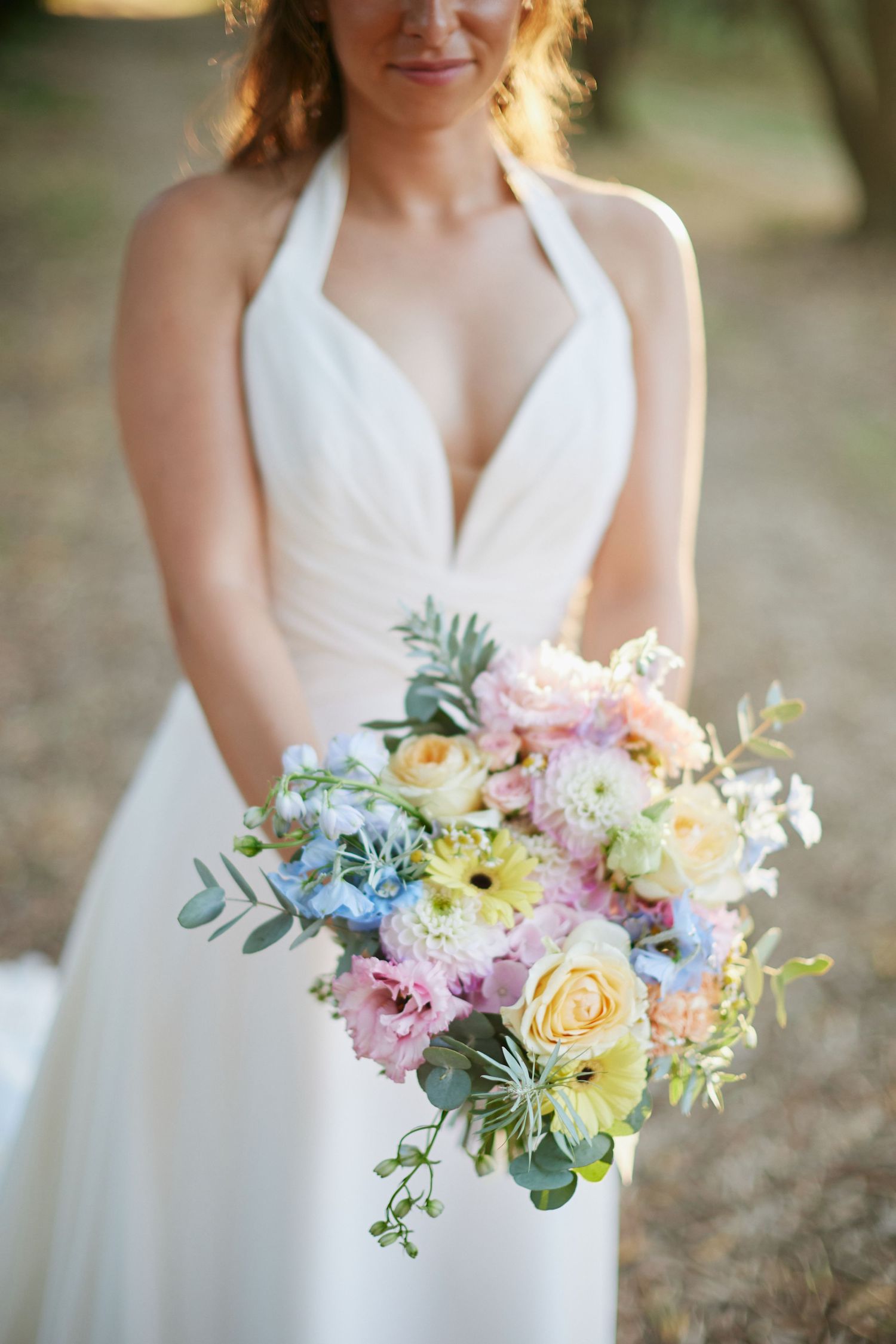 Bride in white halter gown holding a pastel bouquet of roses, hydrangeas, and mixed blooms outdoors.