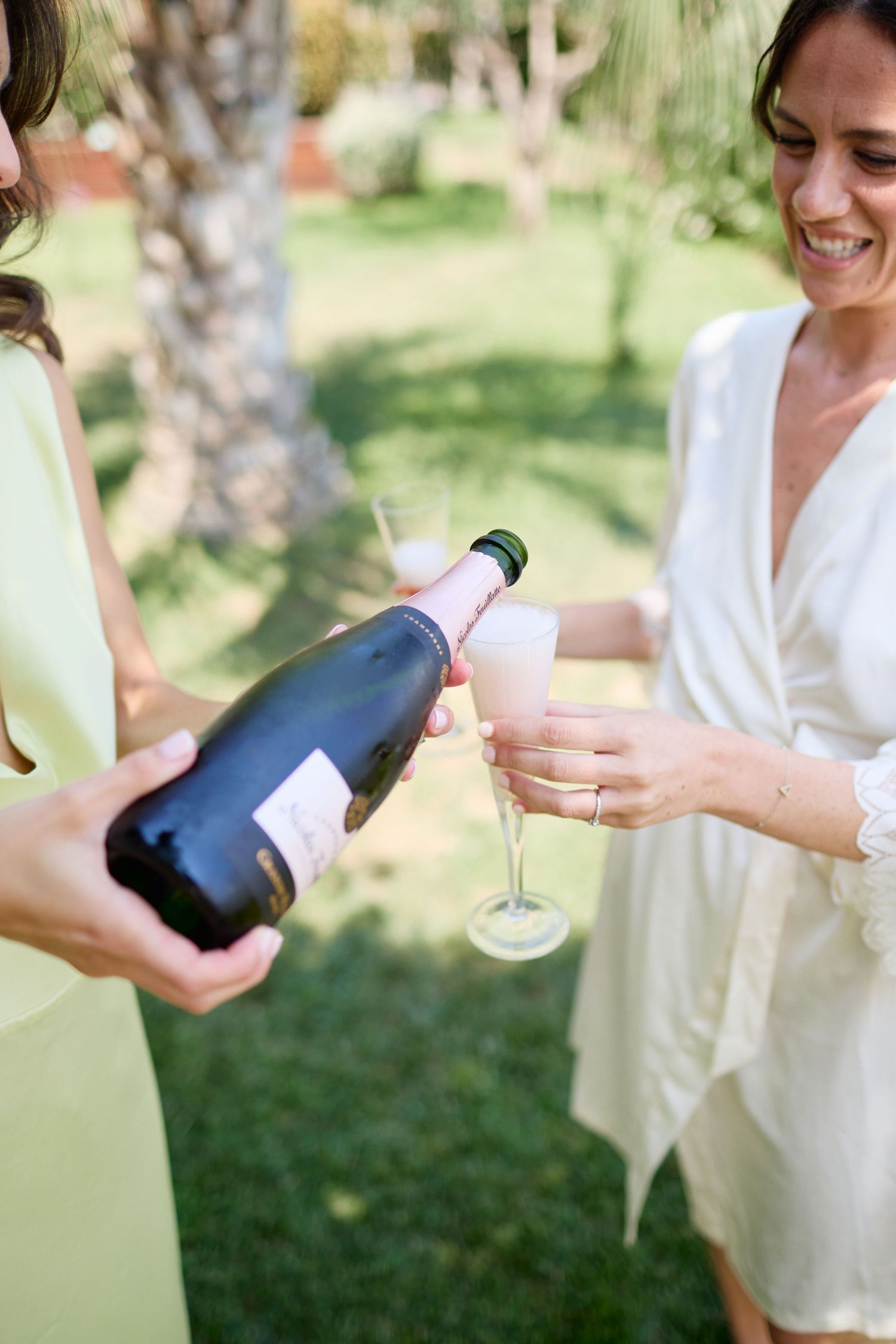 Bridesmaid in yellow dress pouring champagne into flute held by bride in white robe outdoors.
