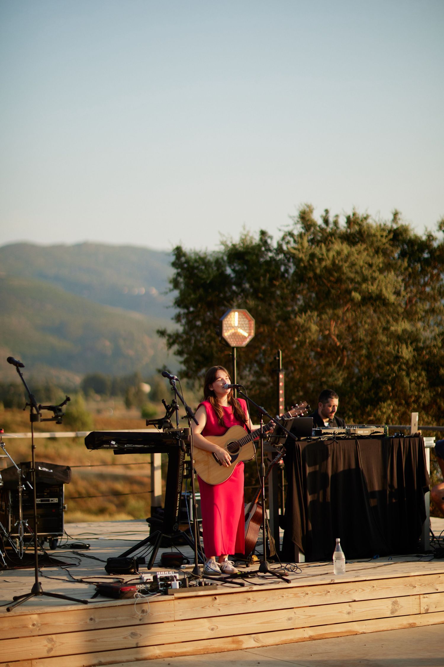 Female musician in red dress performs guitar on outdoor stage at sunset.