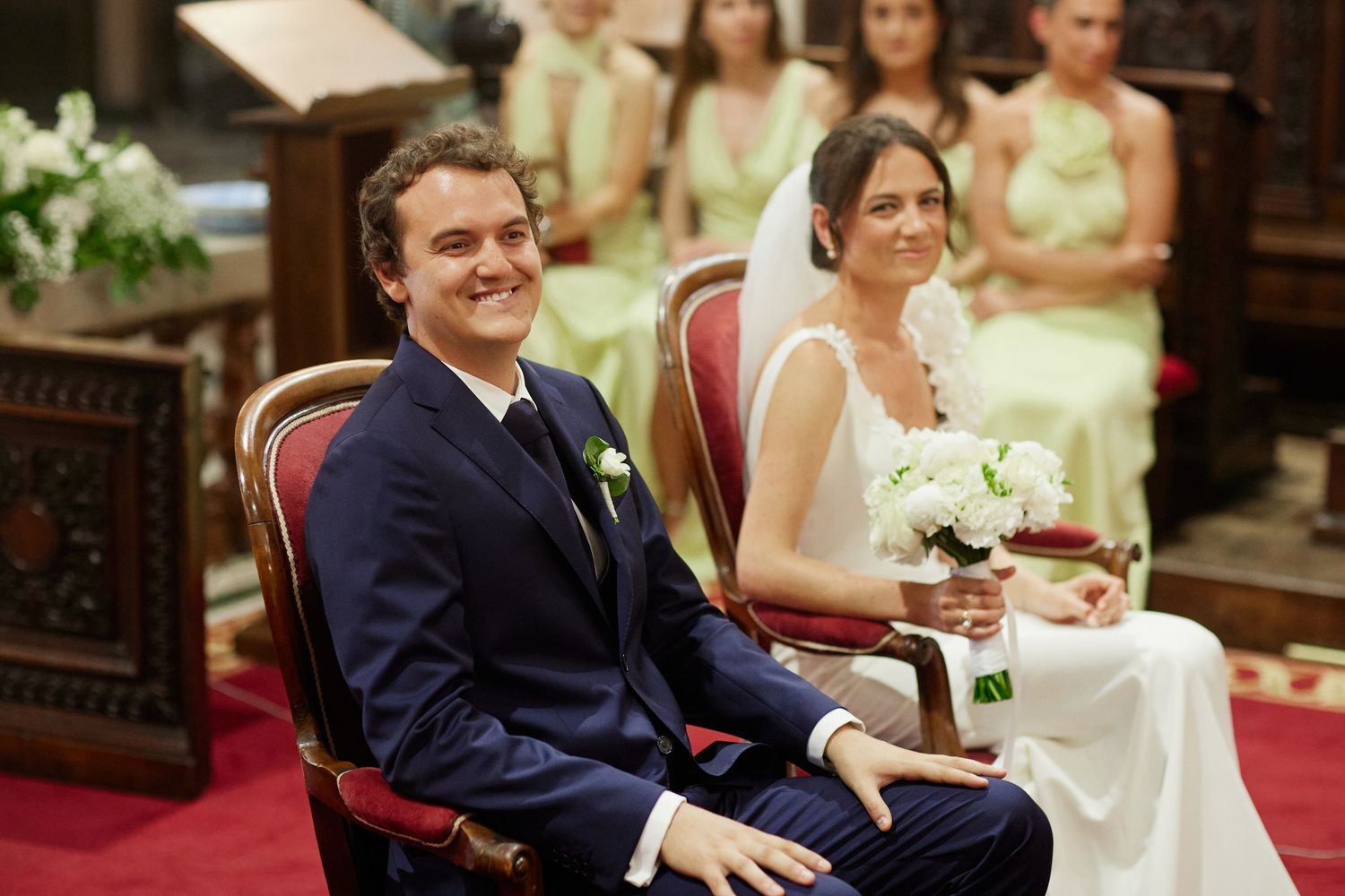 Smiling groom in navy suit seated beside bride holding white bouquet during church ceremony.