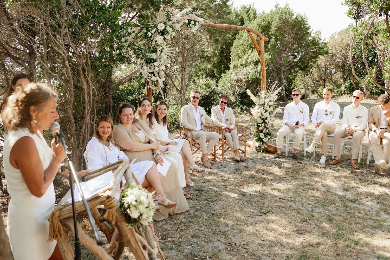 Wedding officiant speaks to seated guests at an elegant outdoor ceremony among lush trees.