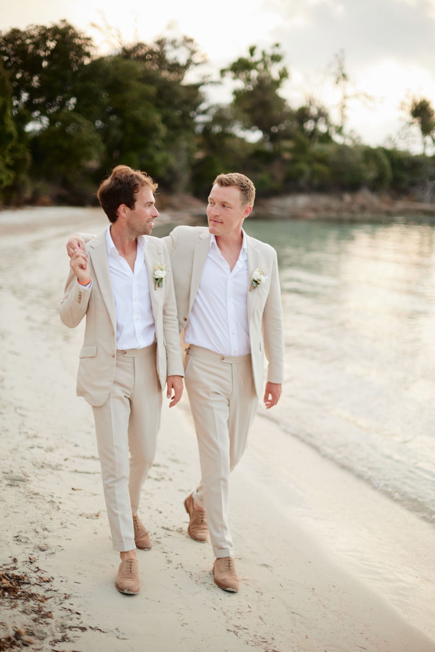 Two grooms in beige linen suits walk arm in arm along a tropical beach at sunset.