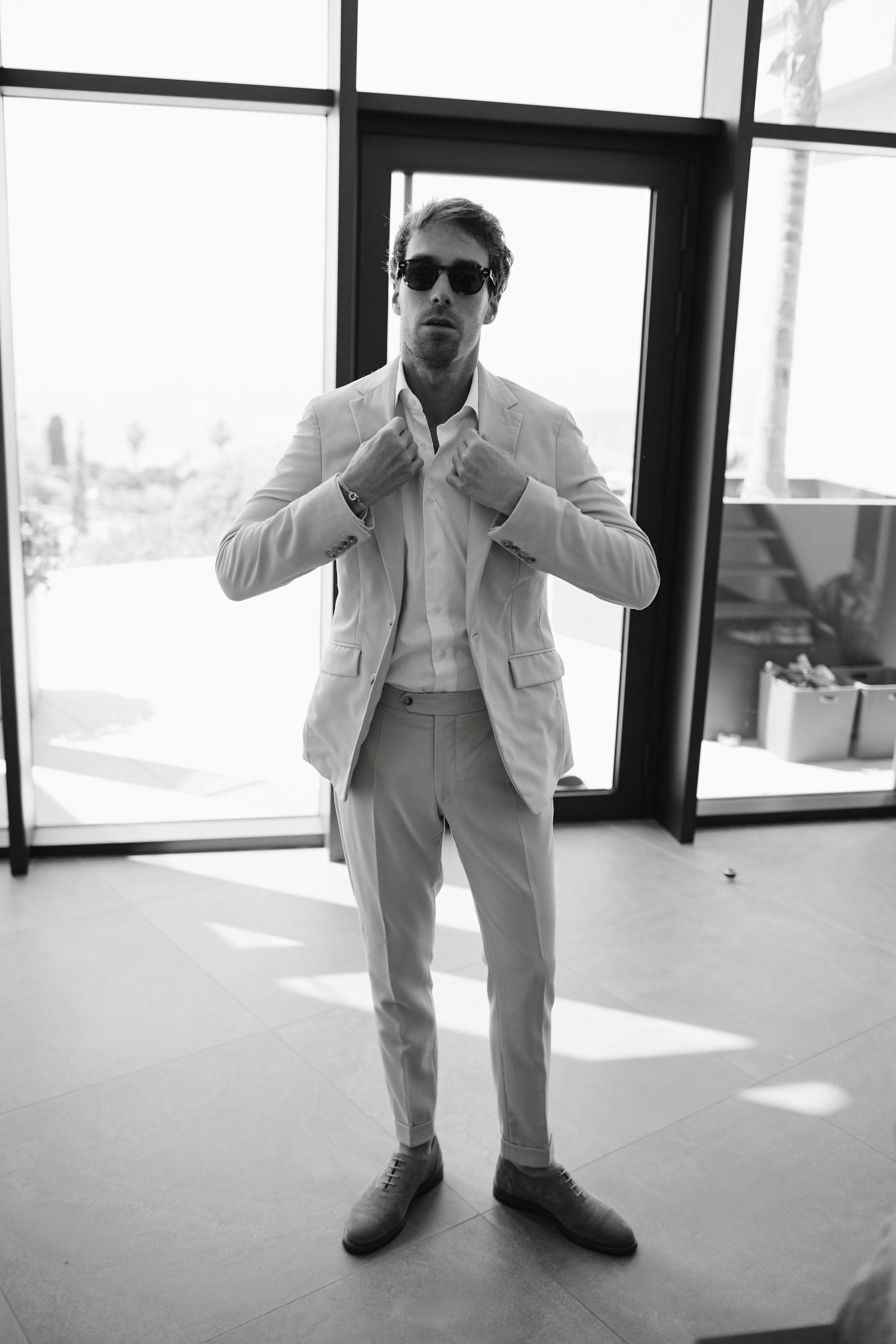 Groom in beige suit and sunglasses adjusting collar in black and white photo by large windows.