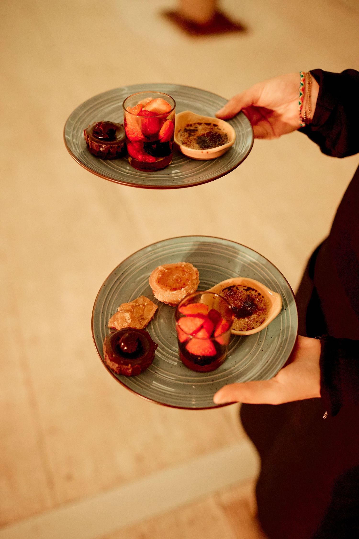 Server holding two elegant plates of assorted desserts including strawberries and creme brulee at a wedding reception.