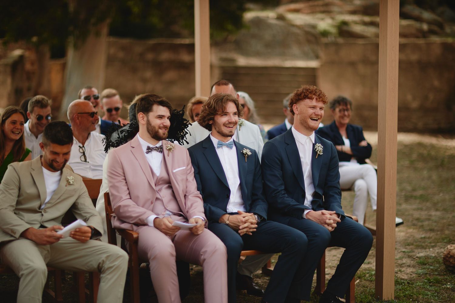 Three groomsmen smiling in pink and navy suits seated at outdoor wedding ceremony with guests behind them.