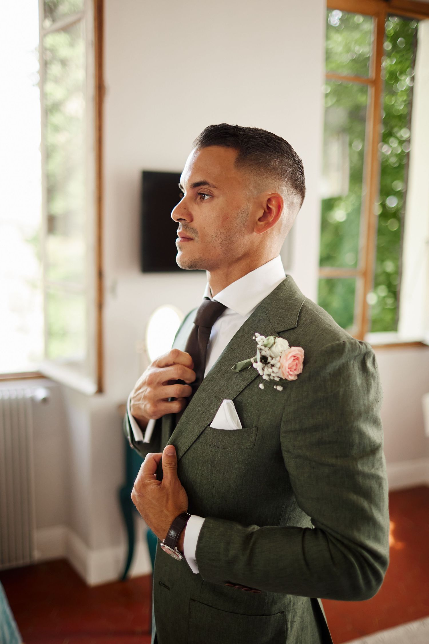 Handsome groom in a green suit adjusting his brown tie with a floral boutonniere by a window.