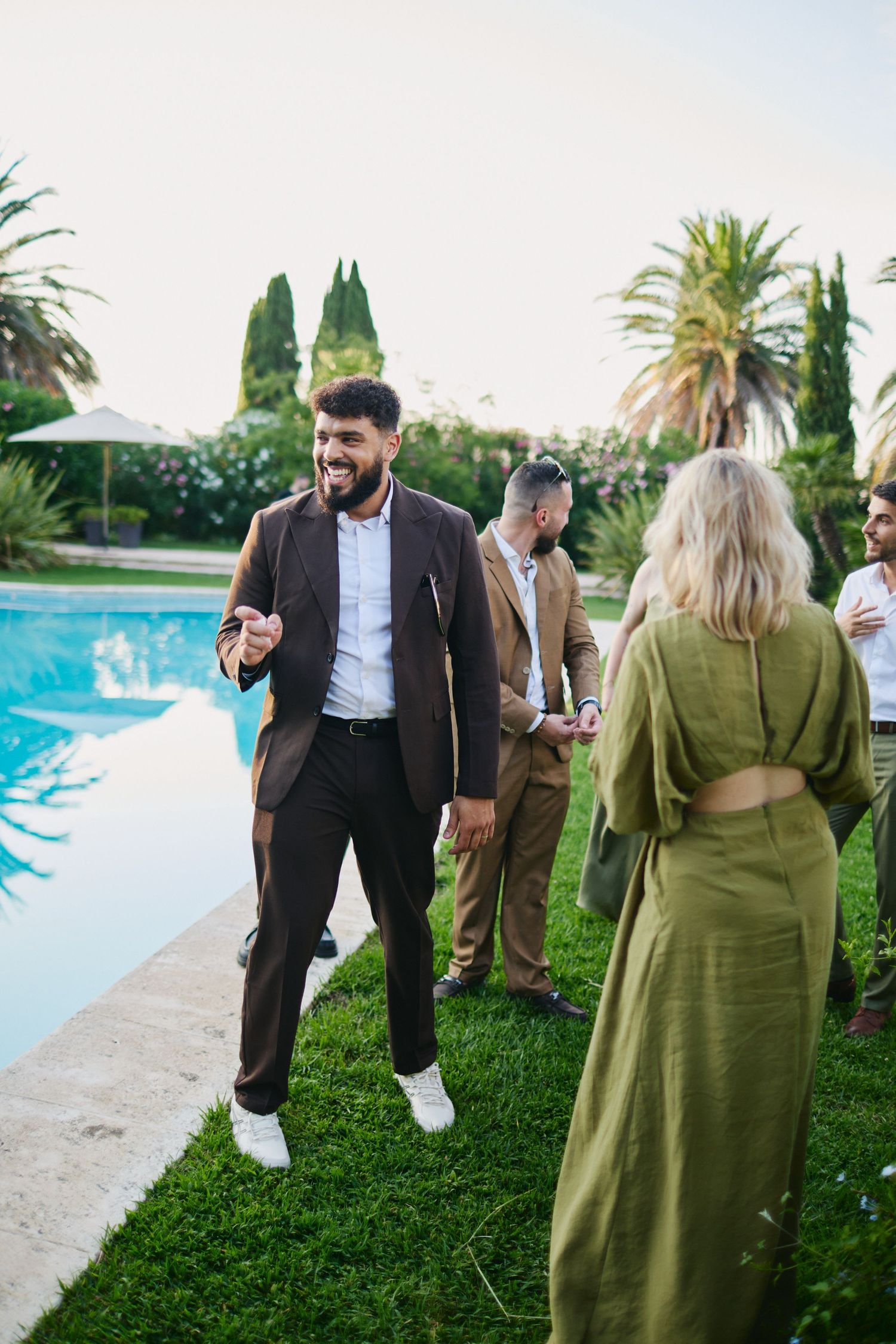 Man in brown suit laughing by poolside at outdoor wedding reception party.