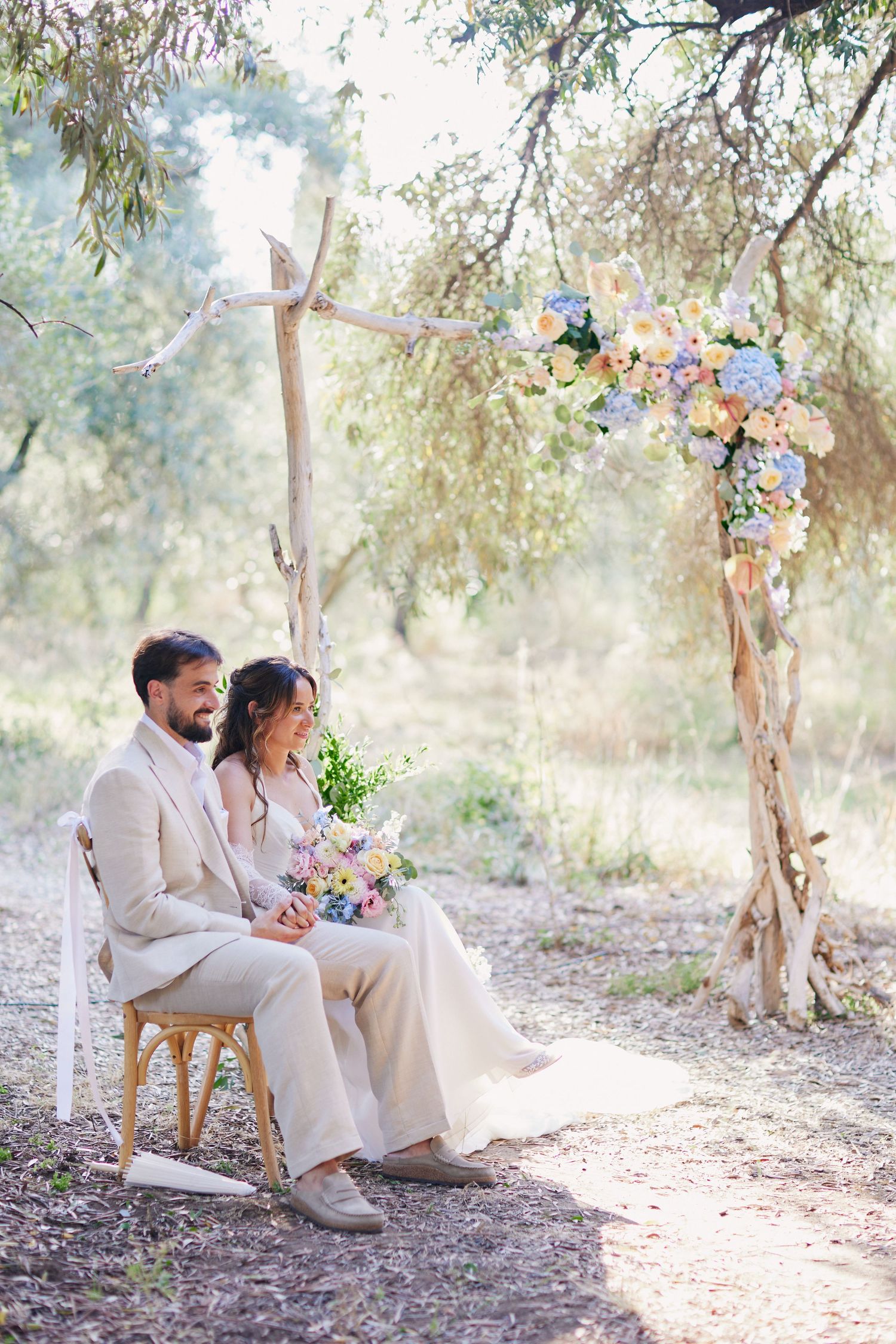 Bride and groom sitting together beneath a rustic wooden floral arch in a sunlit olive grove.