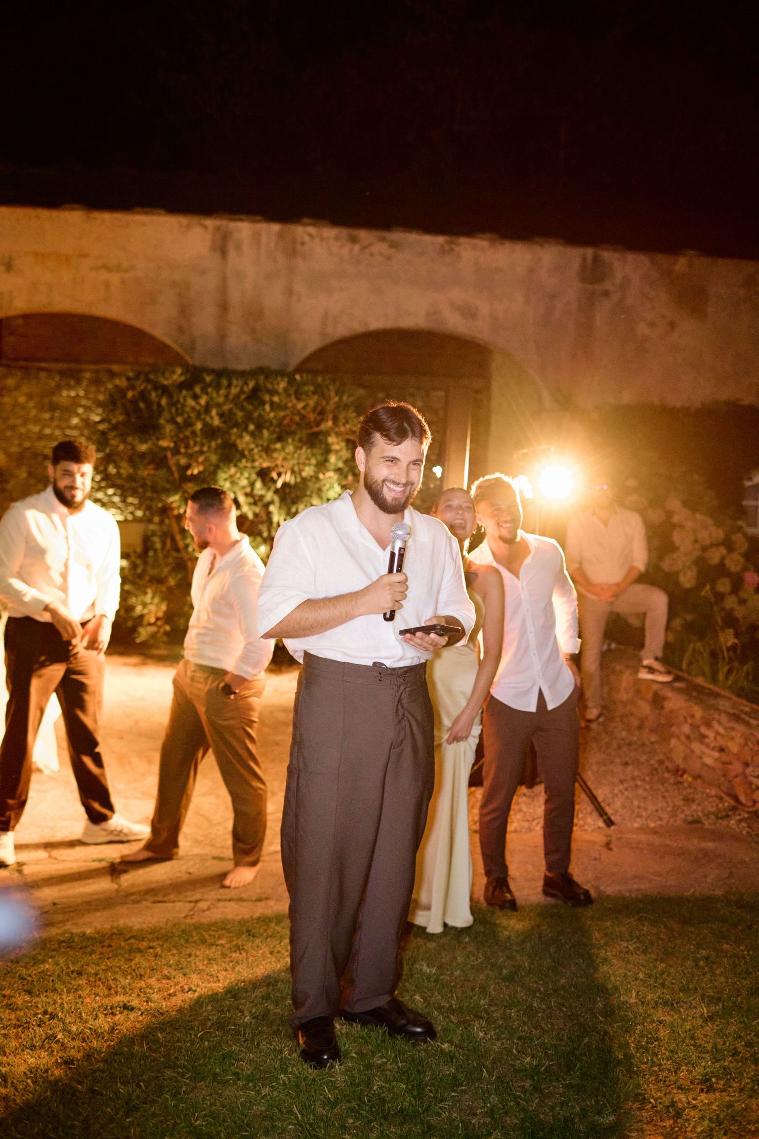 Smiling groomsman holds microphone giving speech at outdoor nighttime wedding reception.