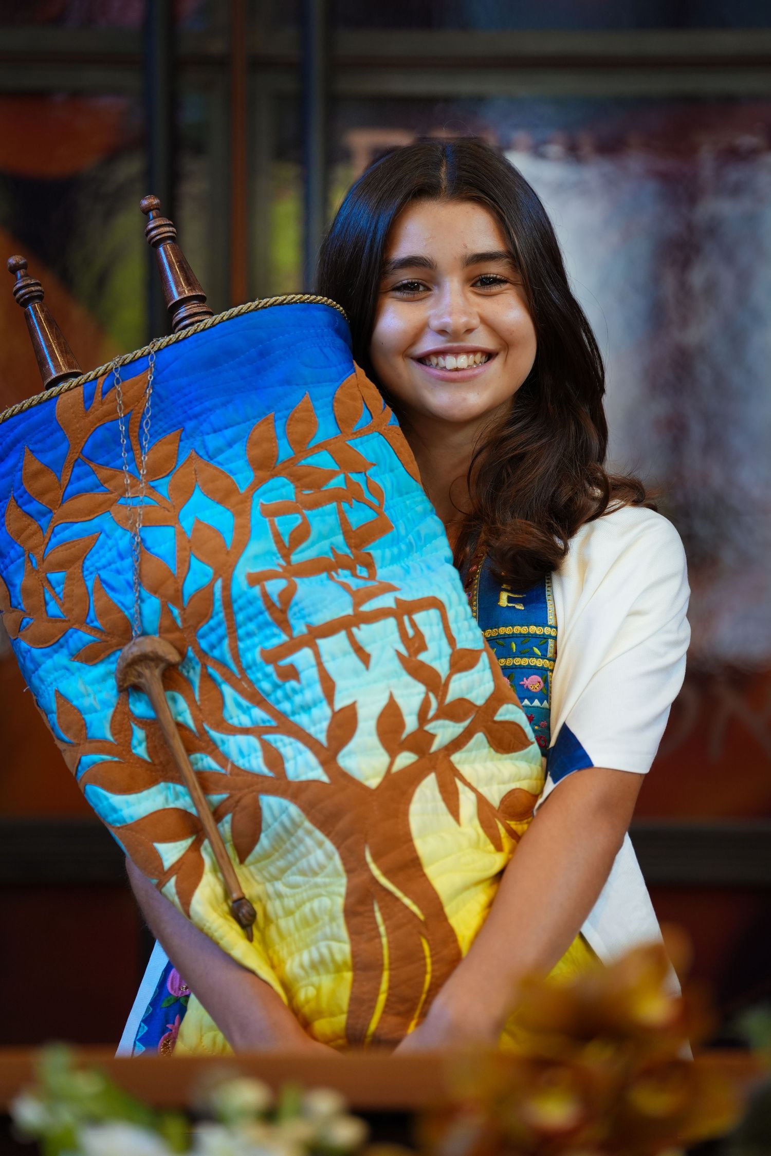 Smiling teenage girl holding a colorful Torah scroll with tree of life design at her Bat Mitzvah ceremony.