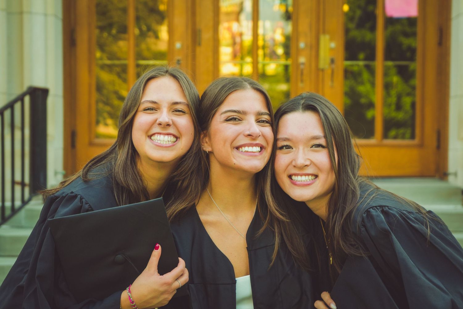 Three smiling women in black graduation gowns pose together outdoors near a building entrance.