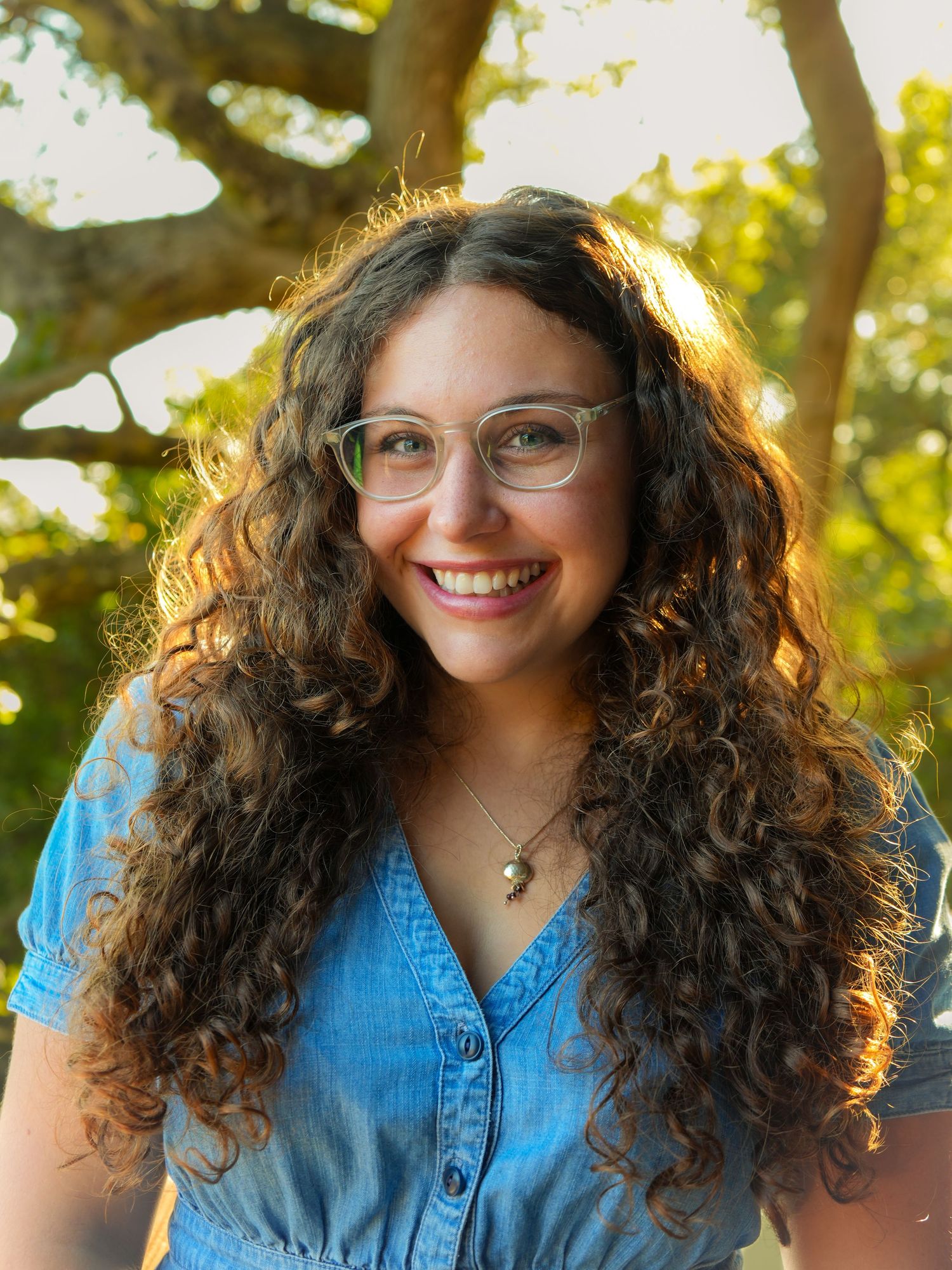 Smiling young woman with curly hair and glasses wearing a blue top outdoors with trees in background.