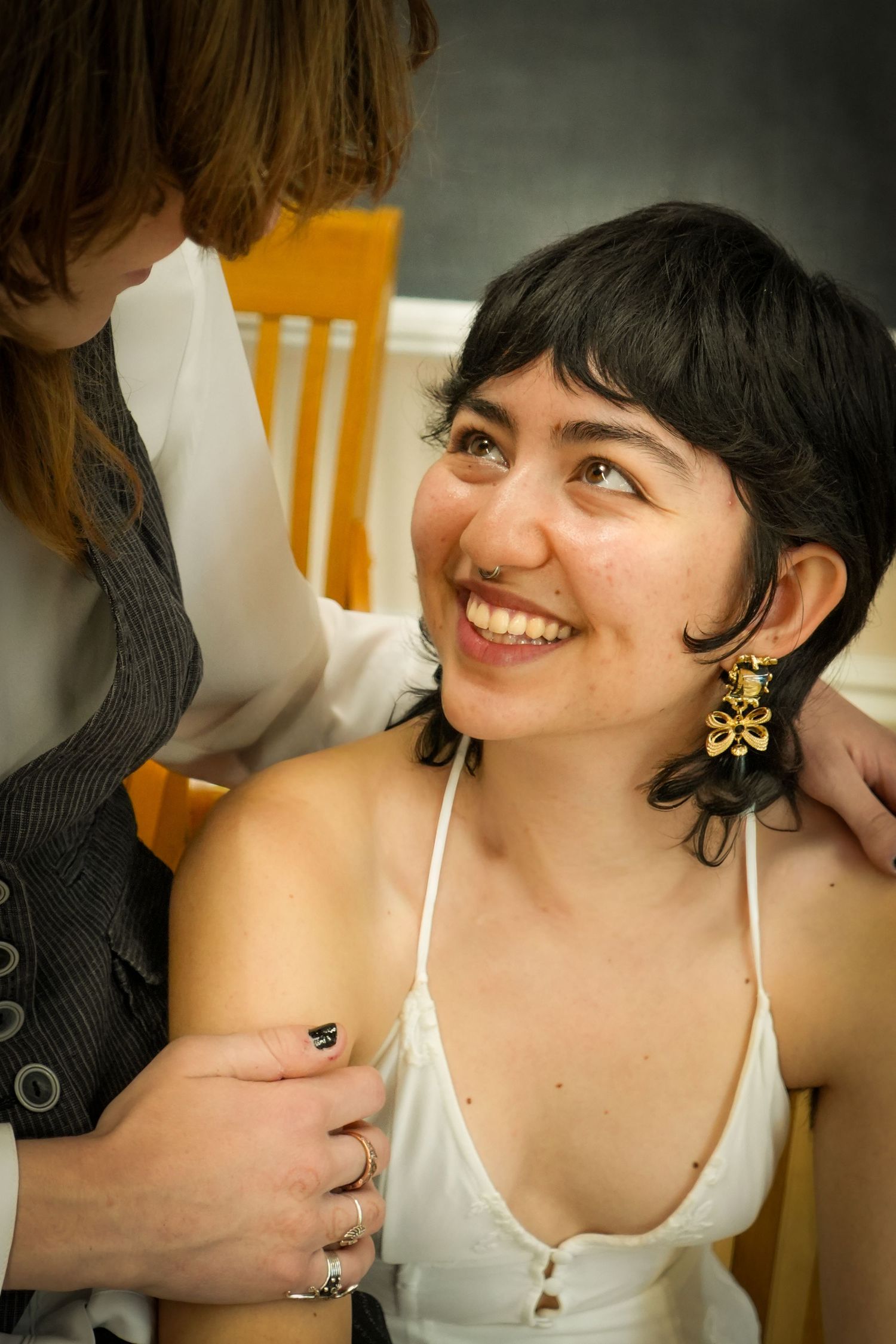 Smiling young woman with dark hair and gold earrings in white top, assisted by another person adjusting her outfit.