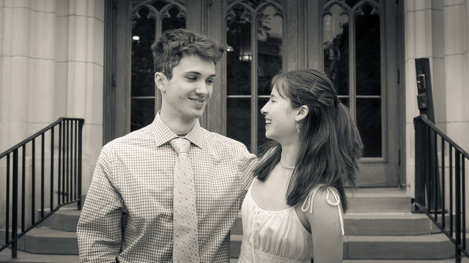 Young couple smiling at each other in black and white photo outside historic building with ornate windows.