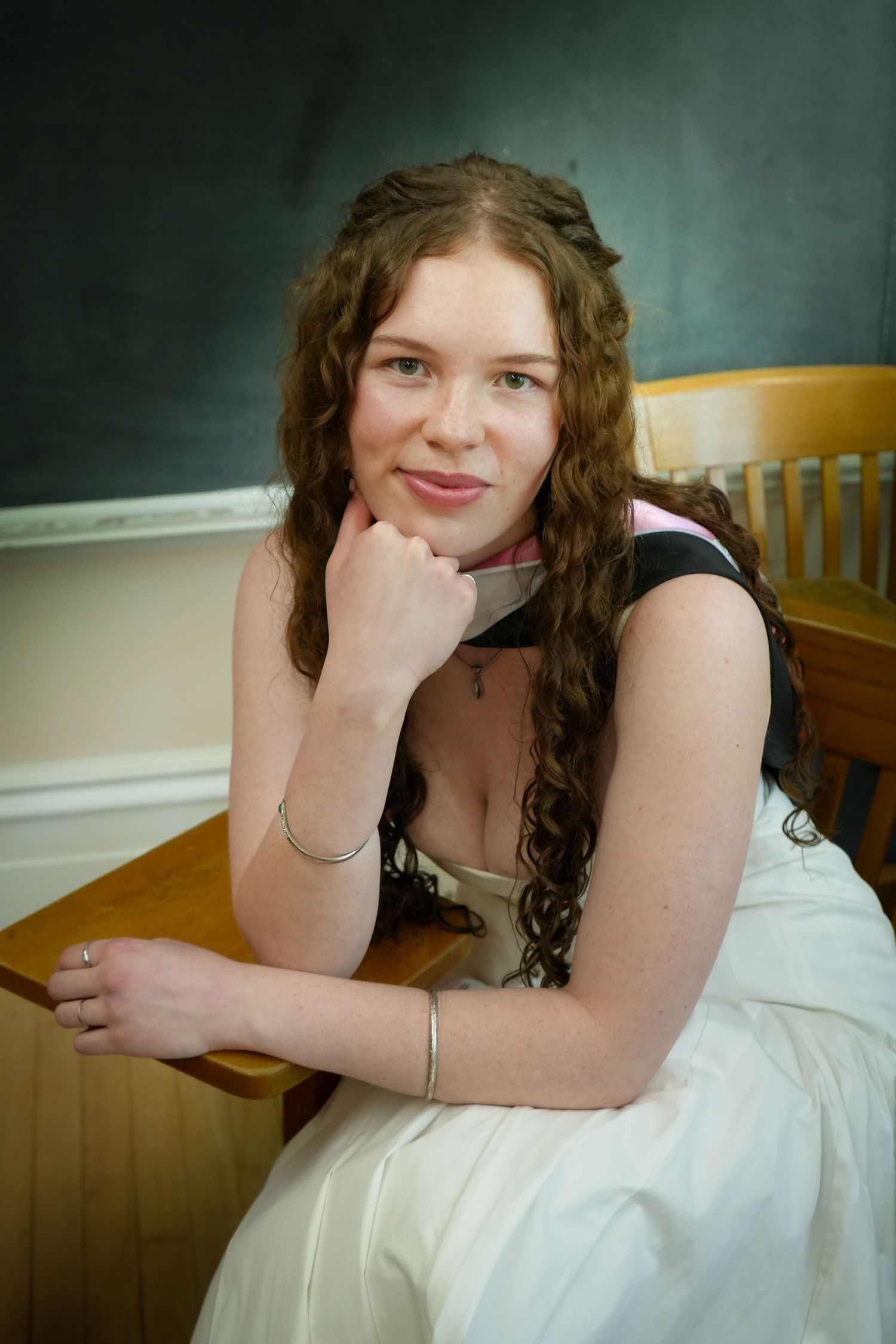 Young woman with curly auburn hair sitting at a wooden school desk, smiling with hand on chin.