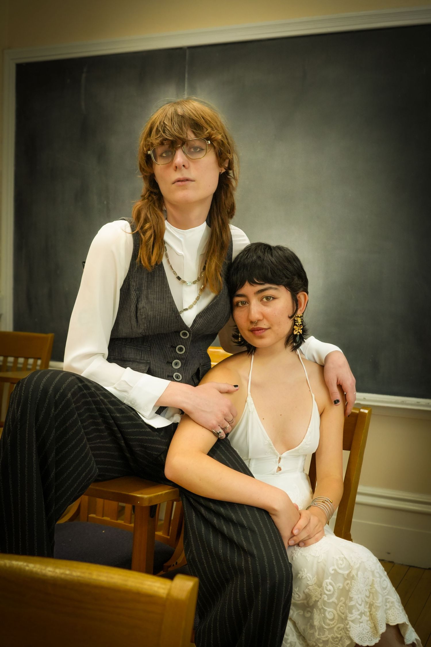 Two people pose formally in front of a chalkboard, one standing in a vest and striped pants, the other seated in a white dress.