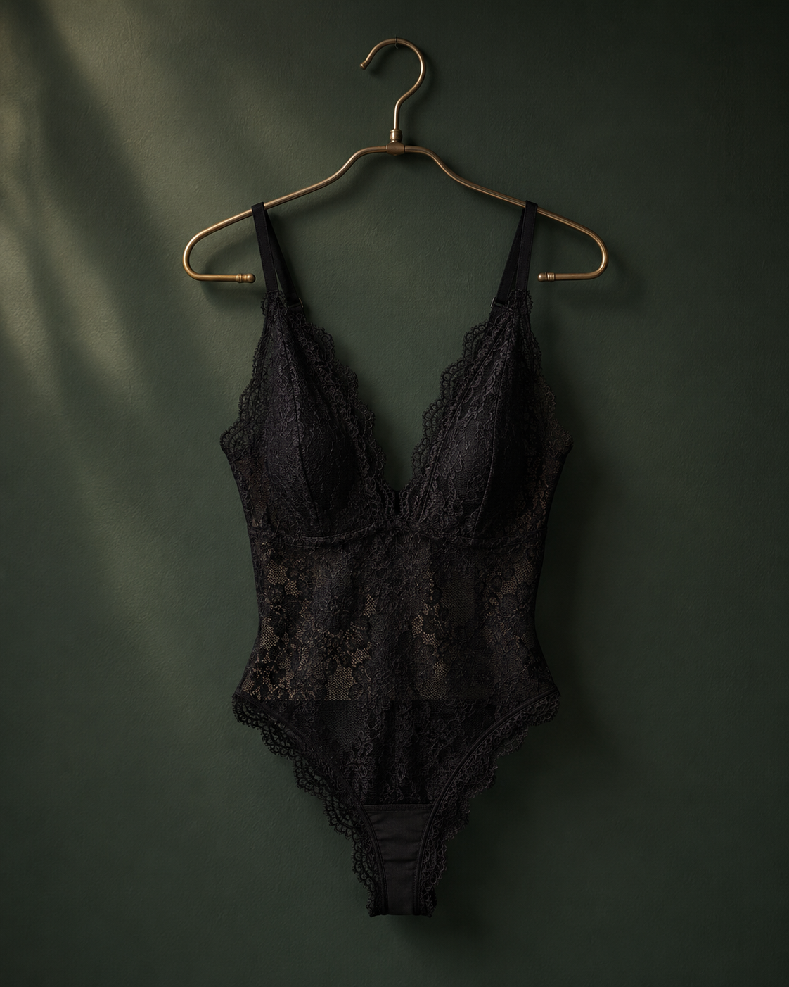 Black floral lace bodysuit displayed on gold hanger against dark green background with soft shadows.