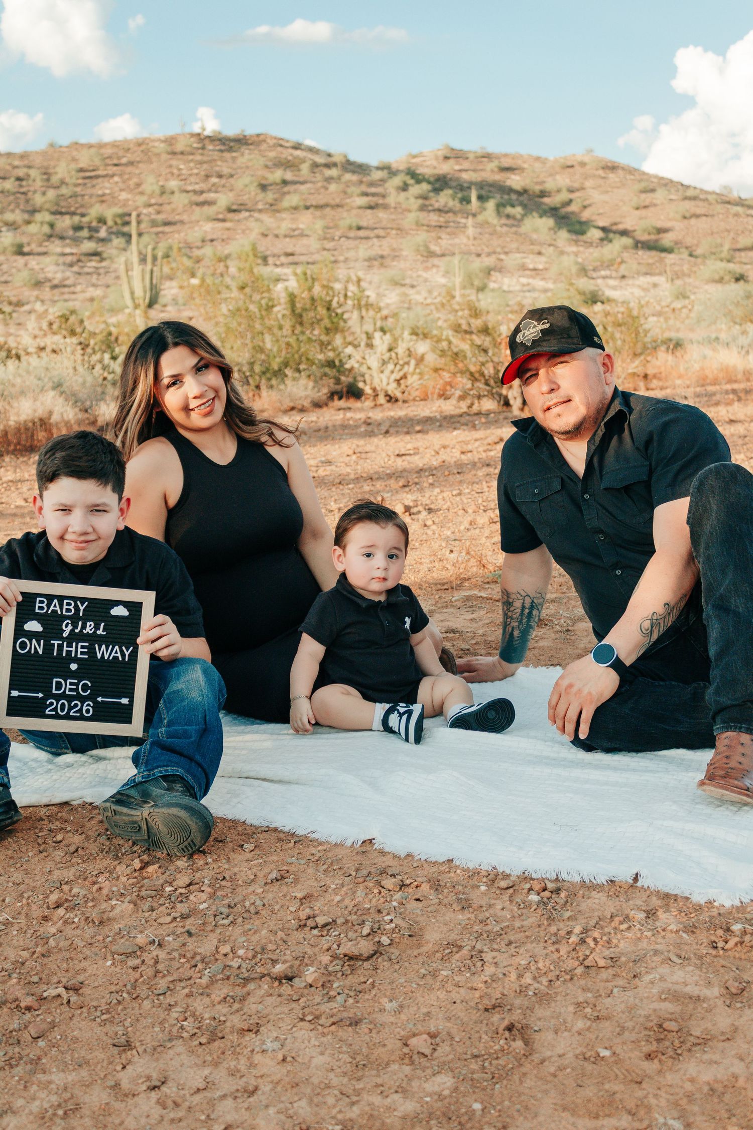 Family pregnancy announcement in desert, holding sign reading Baby Girl On The Way Dec 2026, with two young boys.