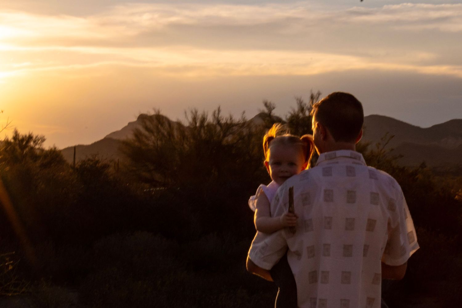Father holding young child watching golden sunset over desert mountains with glowing sky and silhouetted shrubs.
