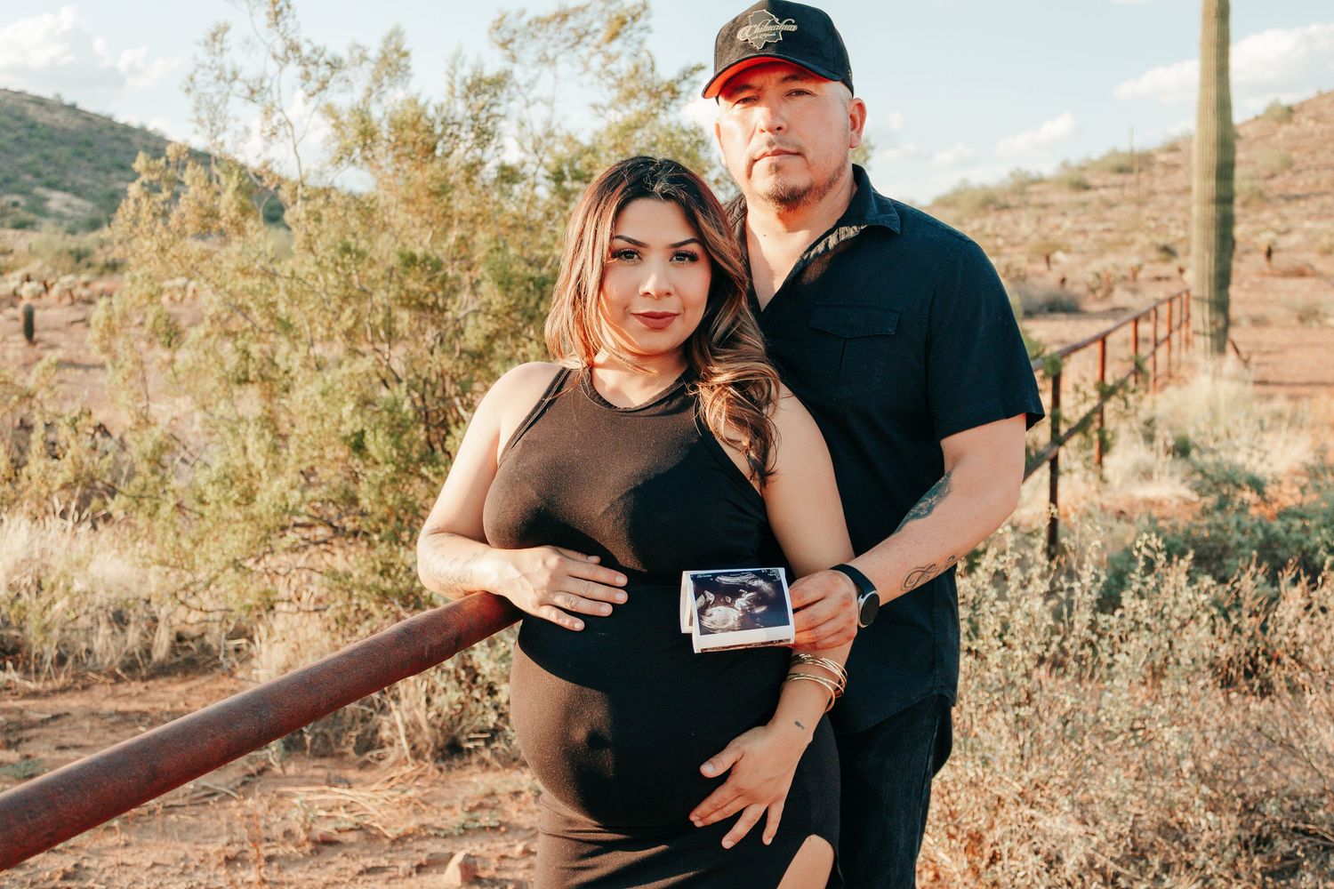 Pregnant couple posing outdoors in desert landscape, holding ultrasound photo, wearing matching black outfits.