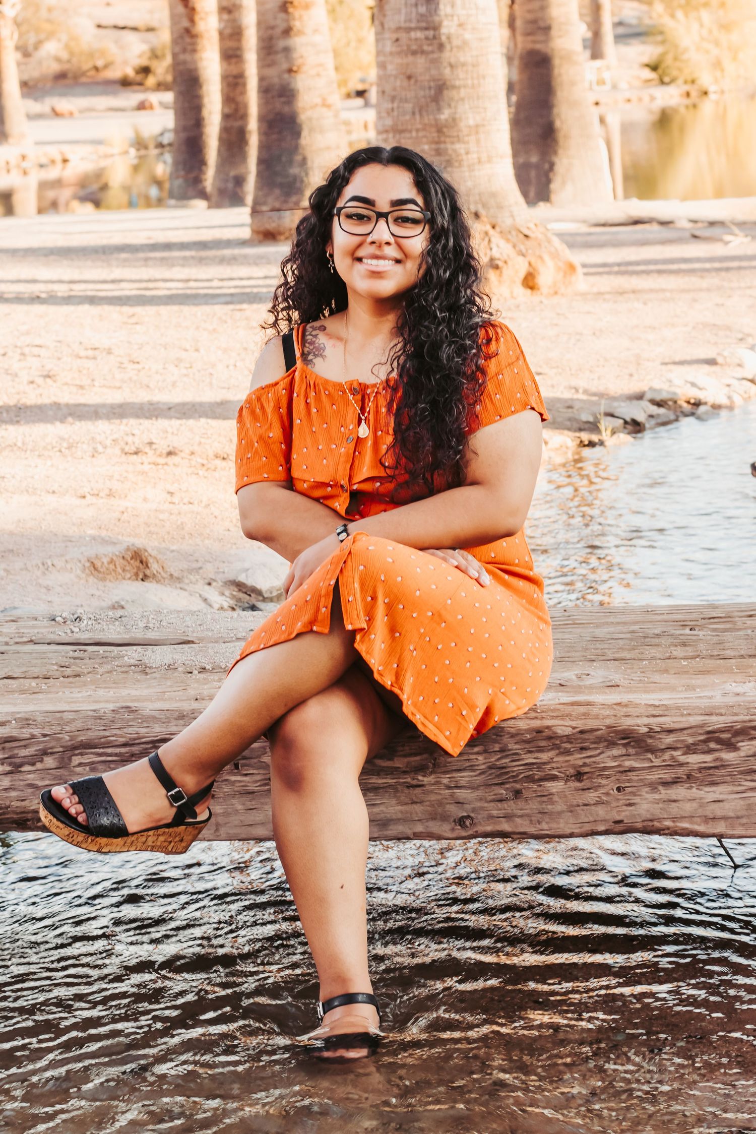 Young woman in orange dress and glasses sitting by water with palm trees, smiling outdoors.