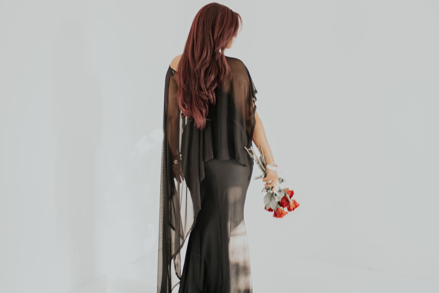 Woman with long red hair in black sheer gown holding red flowers, photographed from behind against white background.