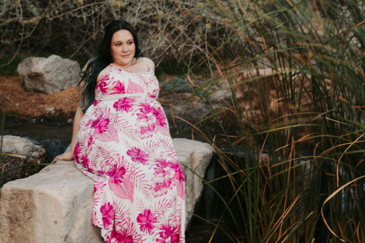 Pregnant woman in pink floral maxi dress sitting on rocks outdoors among tall grasses.